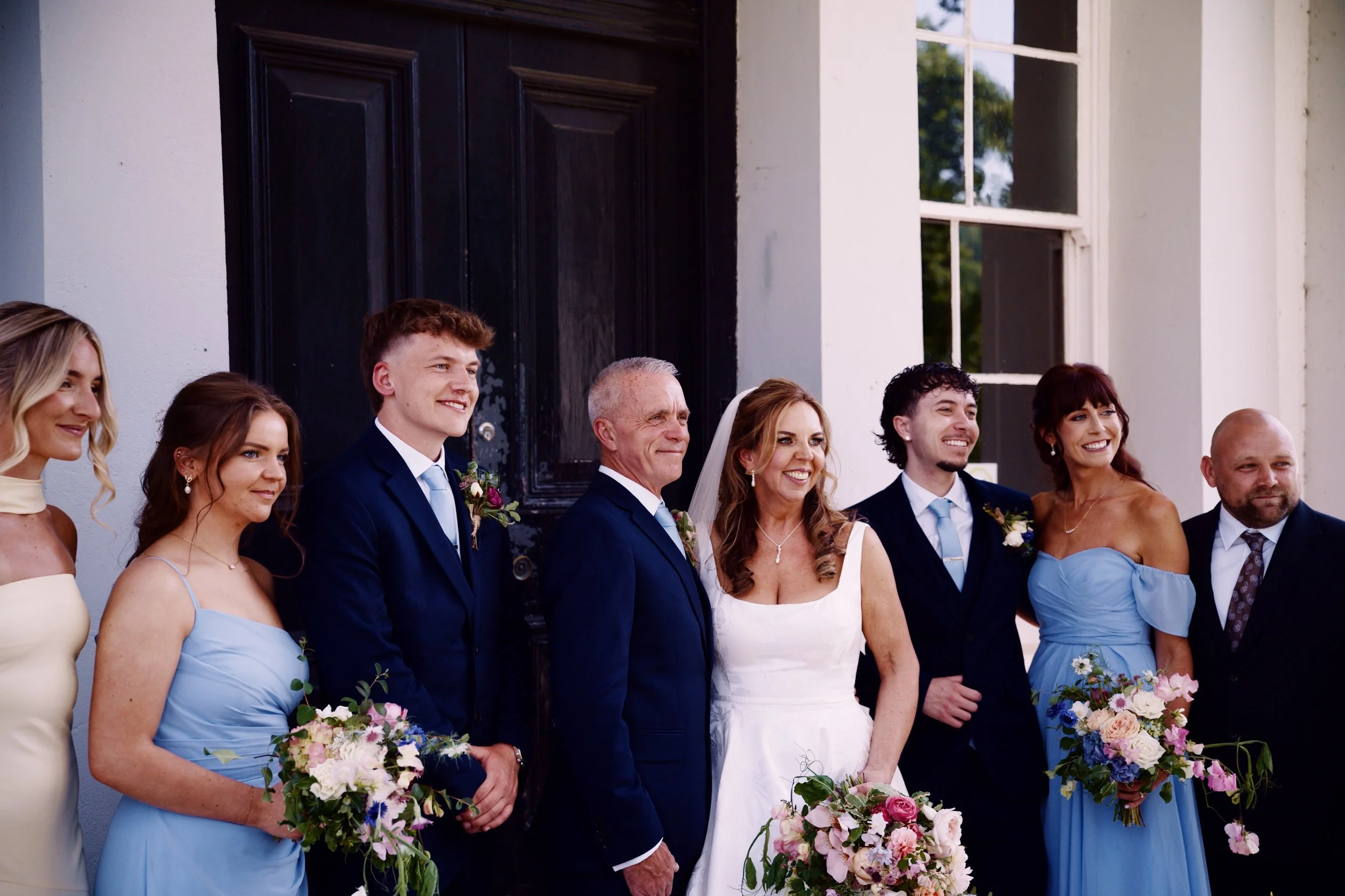 Group of people dressed in formal attire, standing outside in front of a building with large windows, during a wedding celebration.