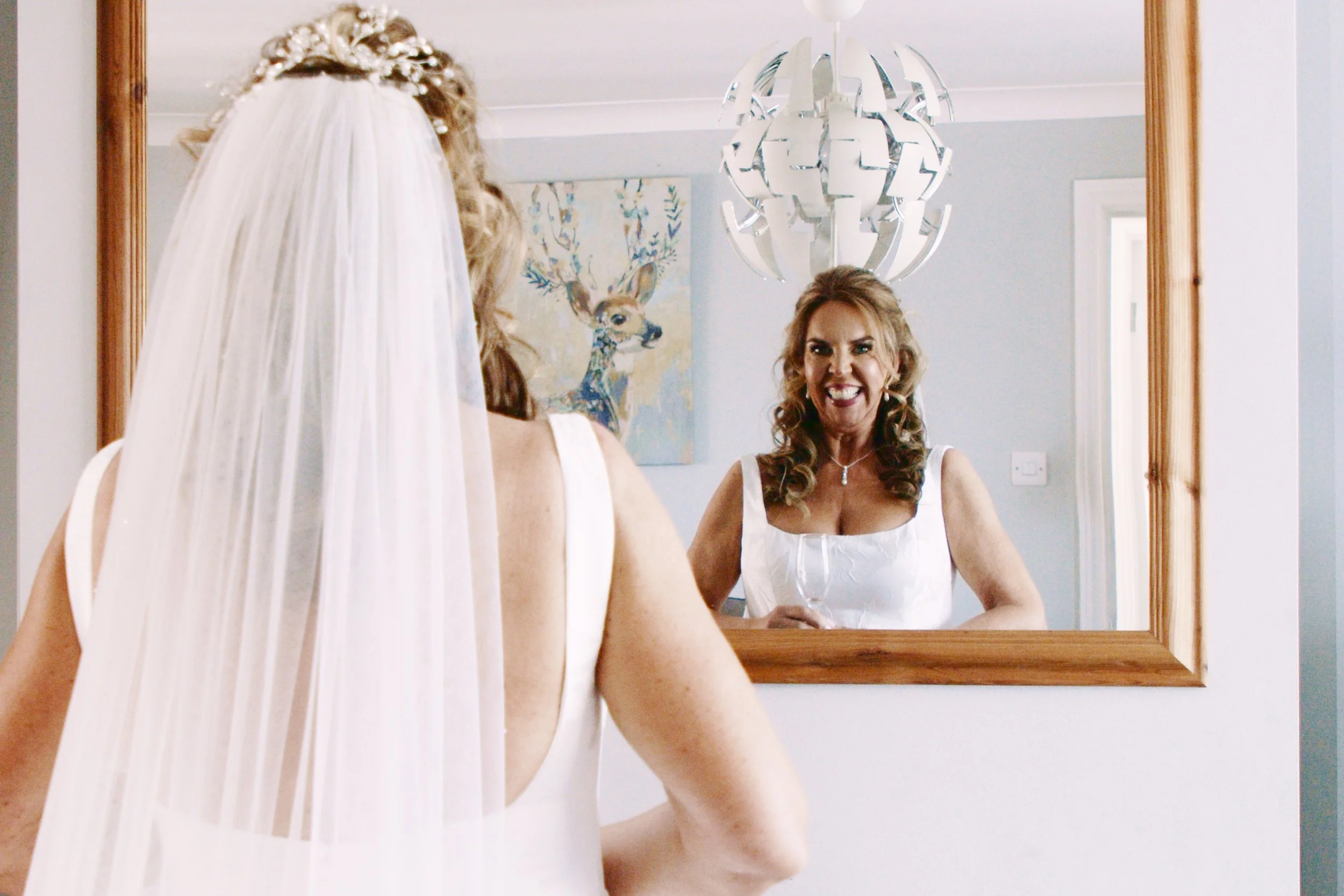 Bride dressed in white dress with veil over her hair smiles in the mirror.