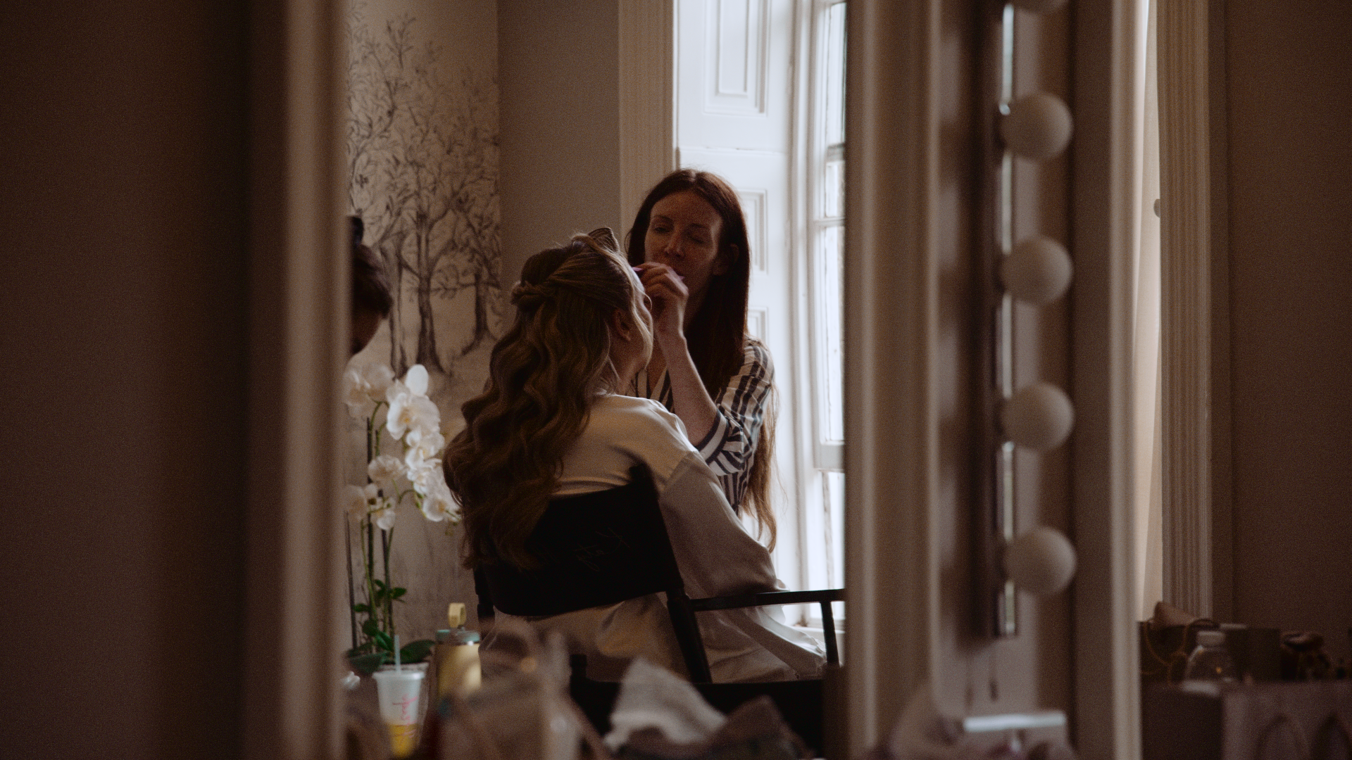 A woman with long hair is applying makeup to a girl with long, light brown hair in a mirror. The scene is framed by a hardware mirror with round bulbs and a window in the background.
