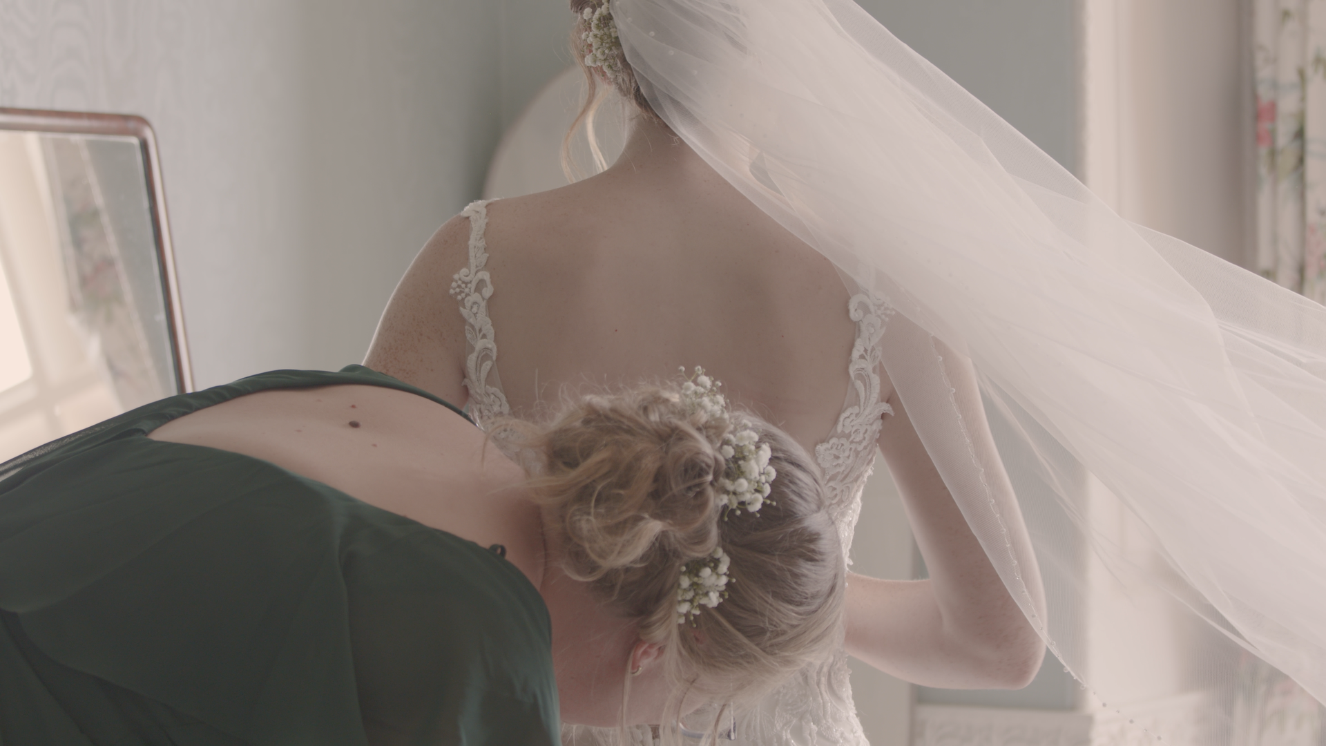 A bride in a lace wedding dress and veil gets ready with a bridesmaid in a dark green dress. The bridesmaid has her head lowered, revealing a floral hairpiece, while soft natural light fills the room.