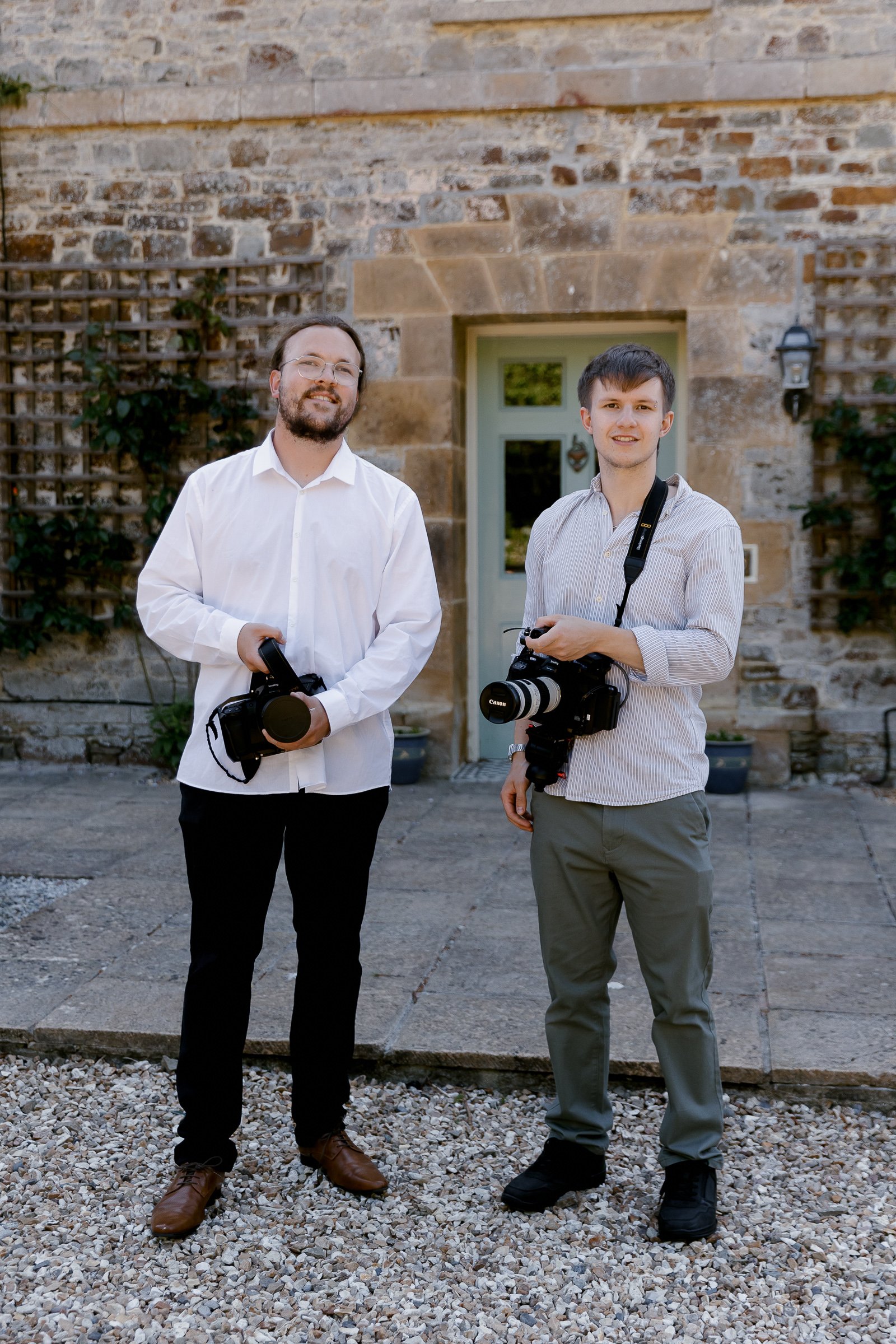 Photograph of the two videographers who make up Littler and Kerti Film. Charlie (left) and Greg (right), they are wearing white formal shirts and dress trousers. Both of them are holding videography cameras.