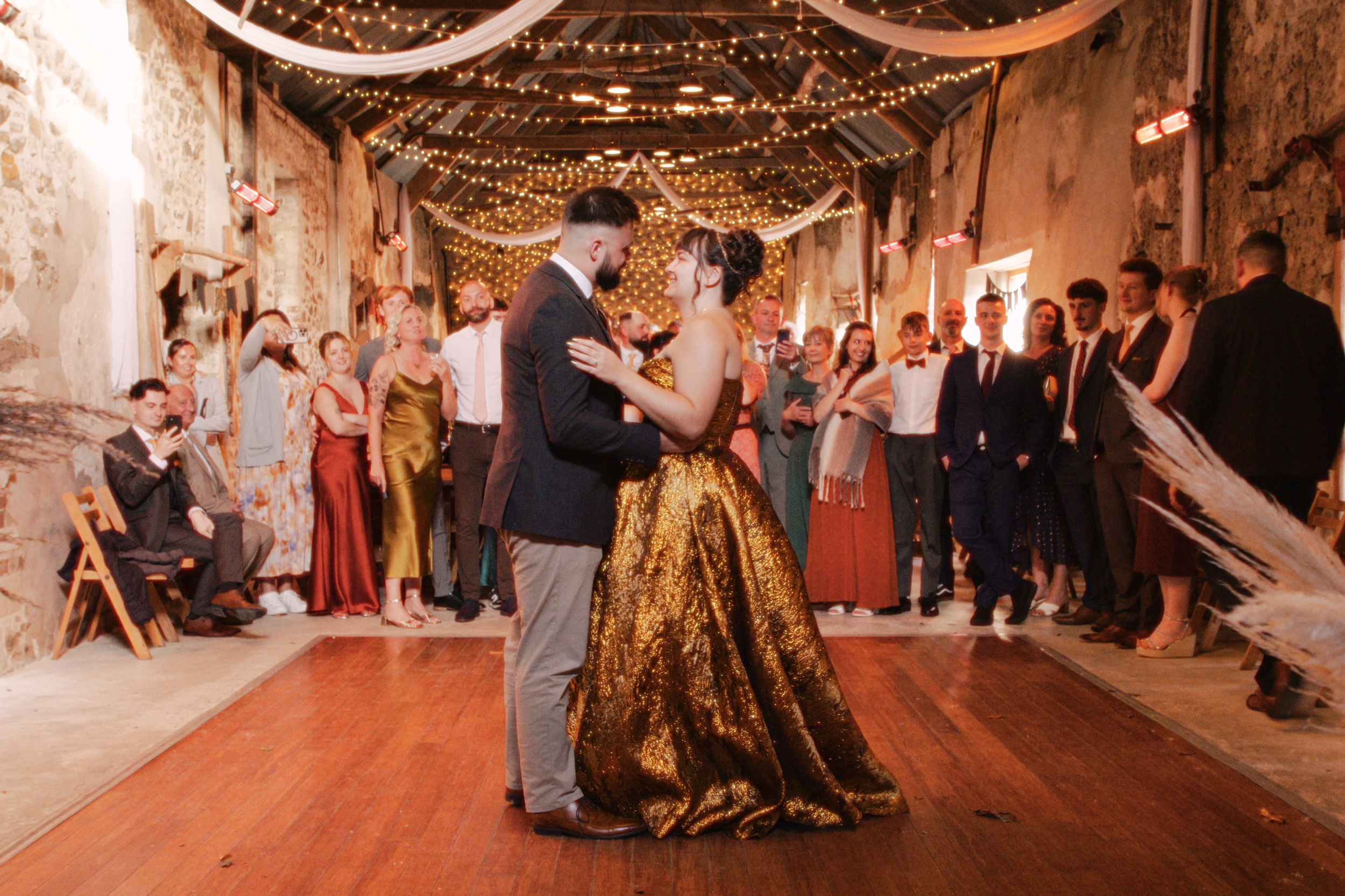Bride and groom have first dance. The groom (left) is wearing a black suit jacket and grey trousers. The bride (right) is wearing a golden floor length wedding dress. In the background is wedding guests watching the couple dance.