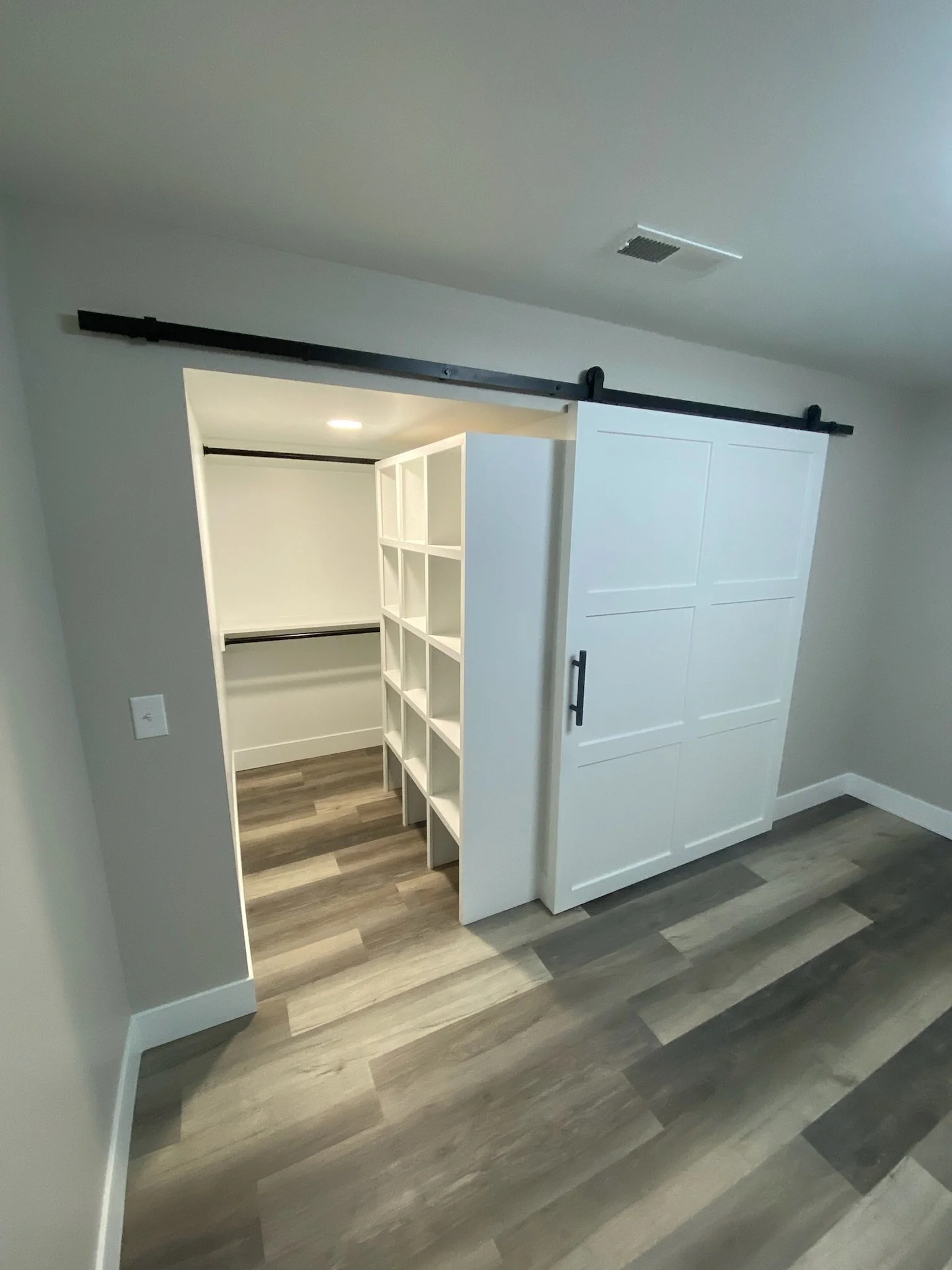 Interior view of an empty walk-in closet with white cabinetry, sliding barn door, black sliding door hardware, gray walls, and wood-look flooring.