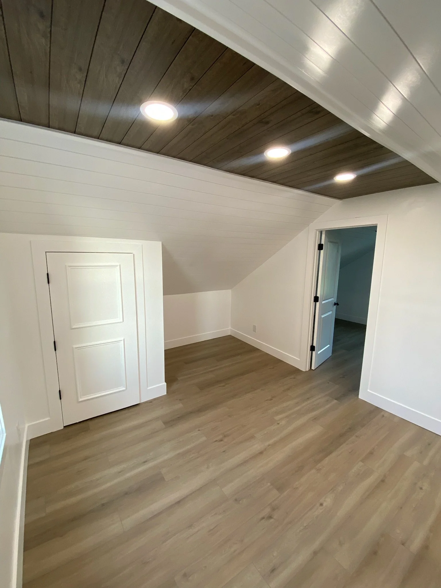 Room with wood flooring, white paneled walls, and a sloped ceiling with dark wood paneling and three recessed lights. There are two small doors, one closed and one open, revealing another room.