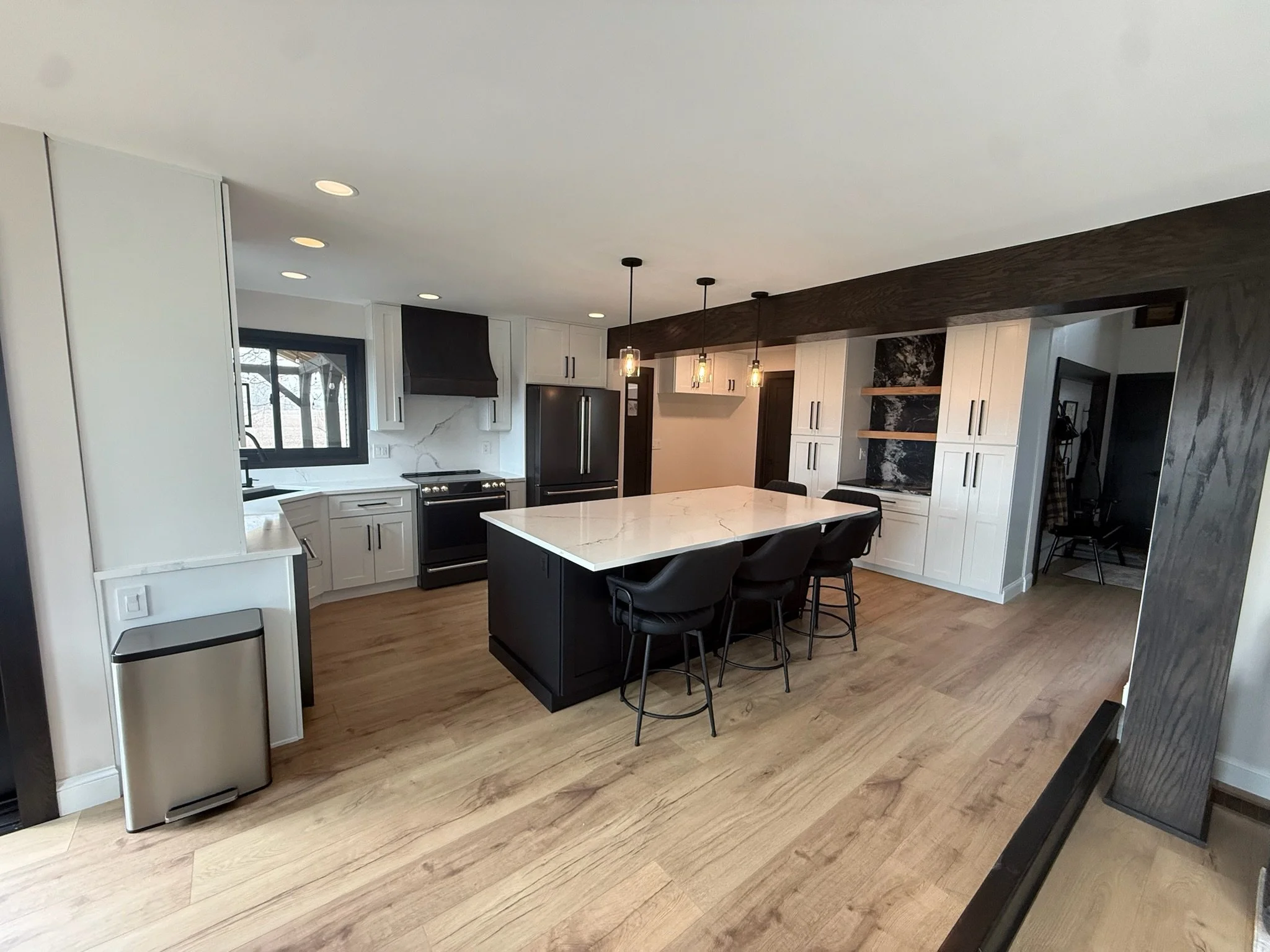 A modern kitchen with white cabinets, black appliances including a refrigerator and stove, a marble island with black bar stools, wooden flooring, pendant lights, a window with natural light, a dark wood beam and a custom hall tree.