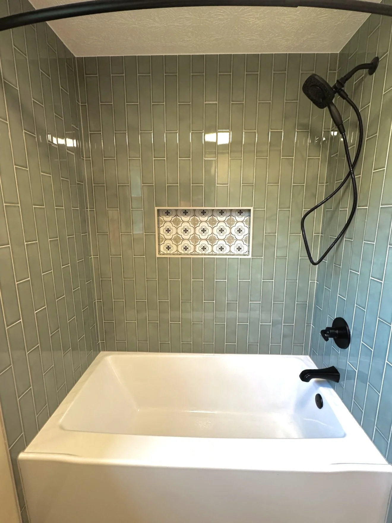 Bathroom shower with green rectangular tile walls, built-in niche with patterned tiles, black showerhead and handles, and a white bathtub at the bottom.