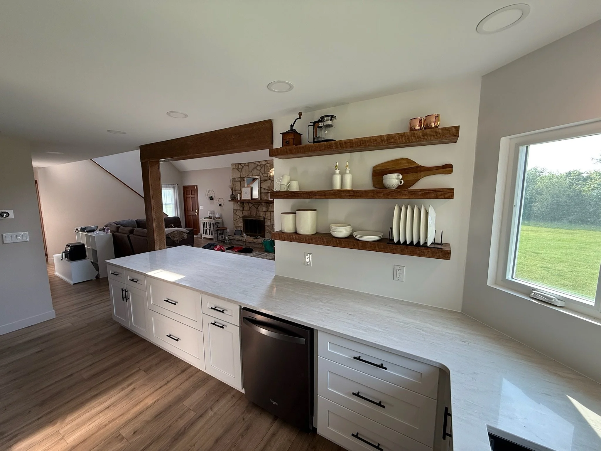 Modern kitchen with white cabinets, a marble countertop, and wooden open shelves displaying dishes and decorative items. Large window with a view of green grass and trees.