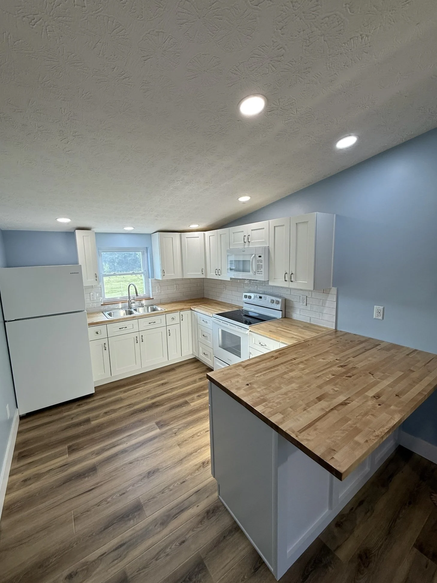Kitchen with white cabinets, butcher block countertops, a white fridge, a microwave above the stove, a sink near the window, and blue walls with a textured ceiling and recessed lighting. Wood-colored flooring. Mother in law suite addition.