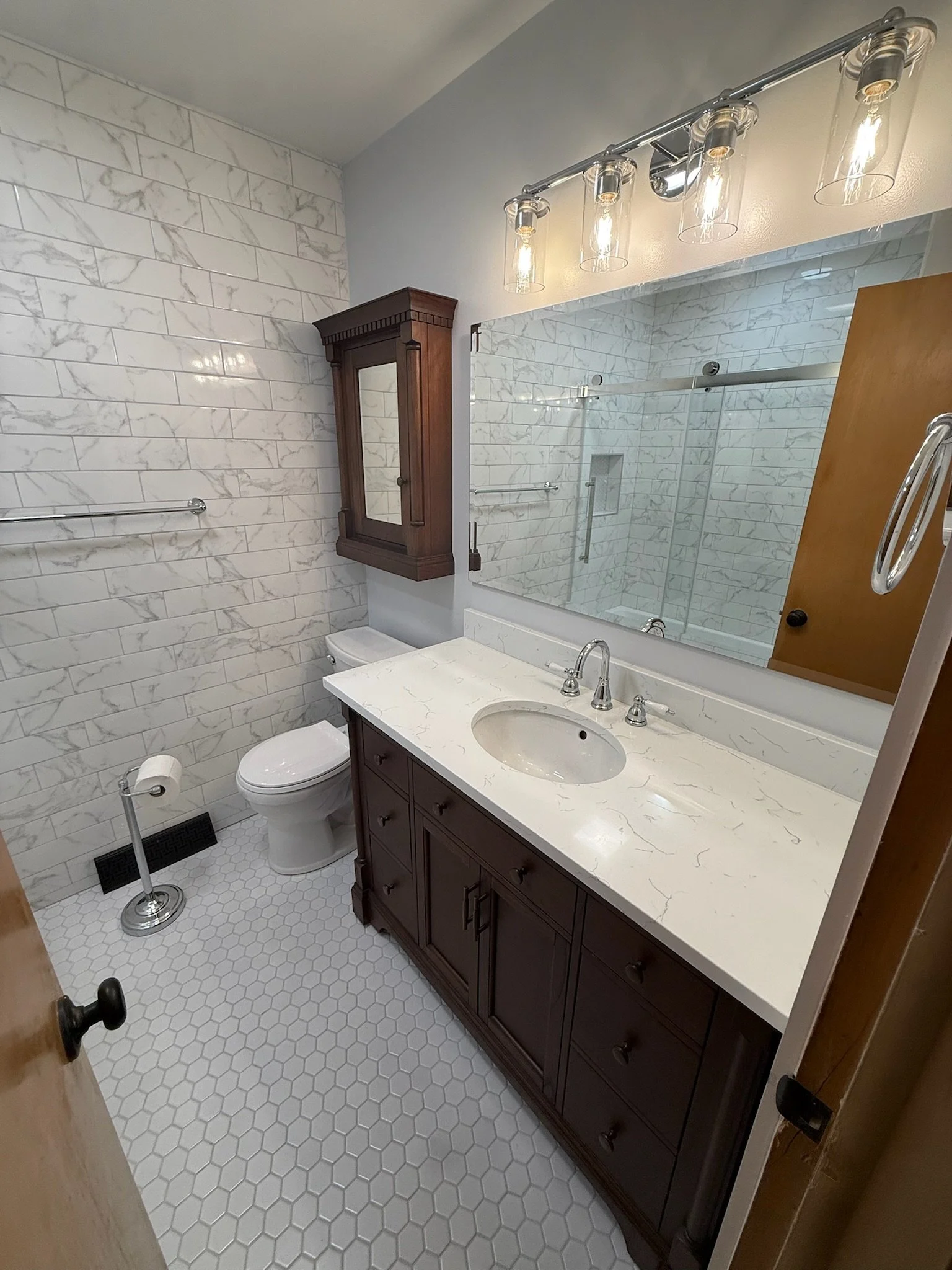 A bathroom with white marble tiled walls and white hexagon tiled floor, featuring a dark wood vanity with a marble countertop, a large mirror, a wooden cabinet with a mirror, a toilet, and a shower area with a glass sliding door.