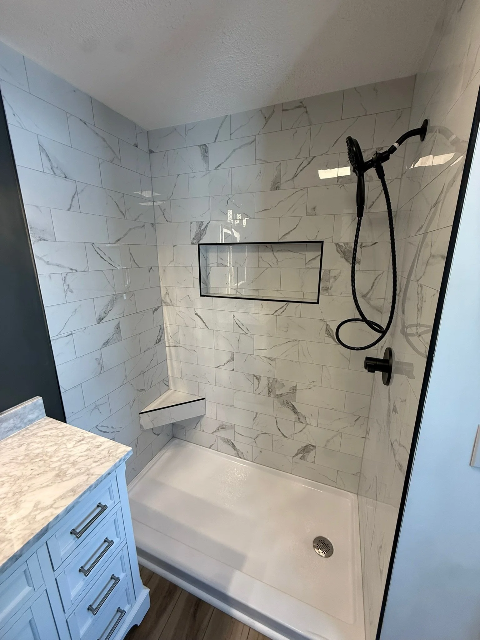A bathroom shower with white marble tiles, a built-in niche, a black showerhead, a blue vanity with a beige marble countertop, and a wood floor.