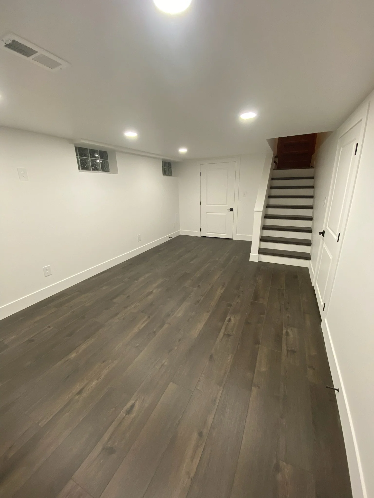 Interior of a basement room with white walls, dark hardwood flooring, a staircase with dark wooden steps, a closed white door, and small glass block windows.