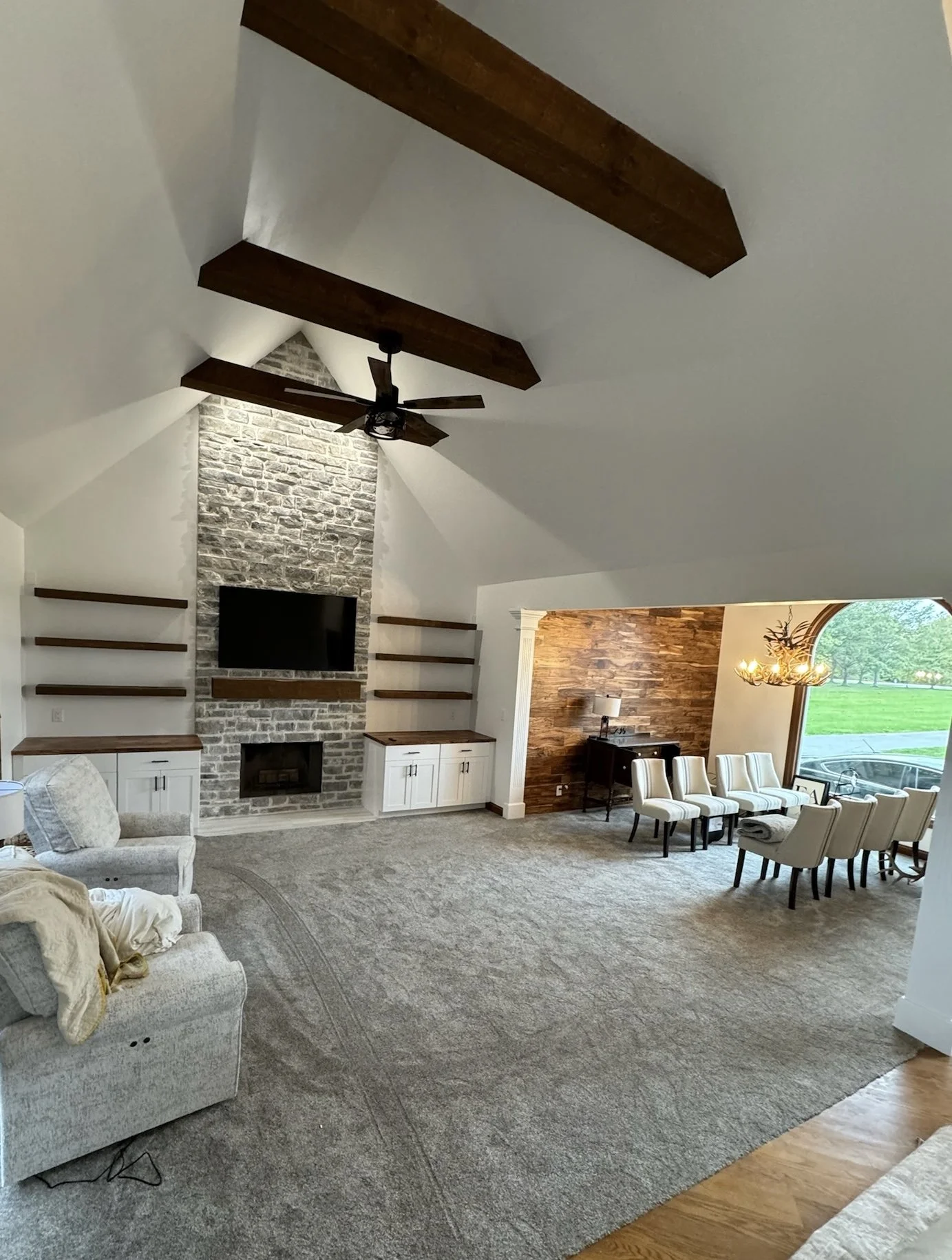 Living room with vaulted ceiling and exposed wooden beams, stone fireplace with mounted TV, white built-in cabinets, beige carpet, beige armchair, and a dining area with white chairs and a wooden wall, large window showing outside greenery.