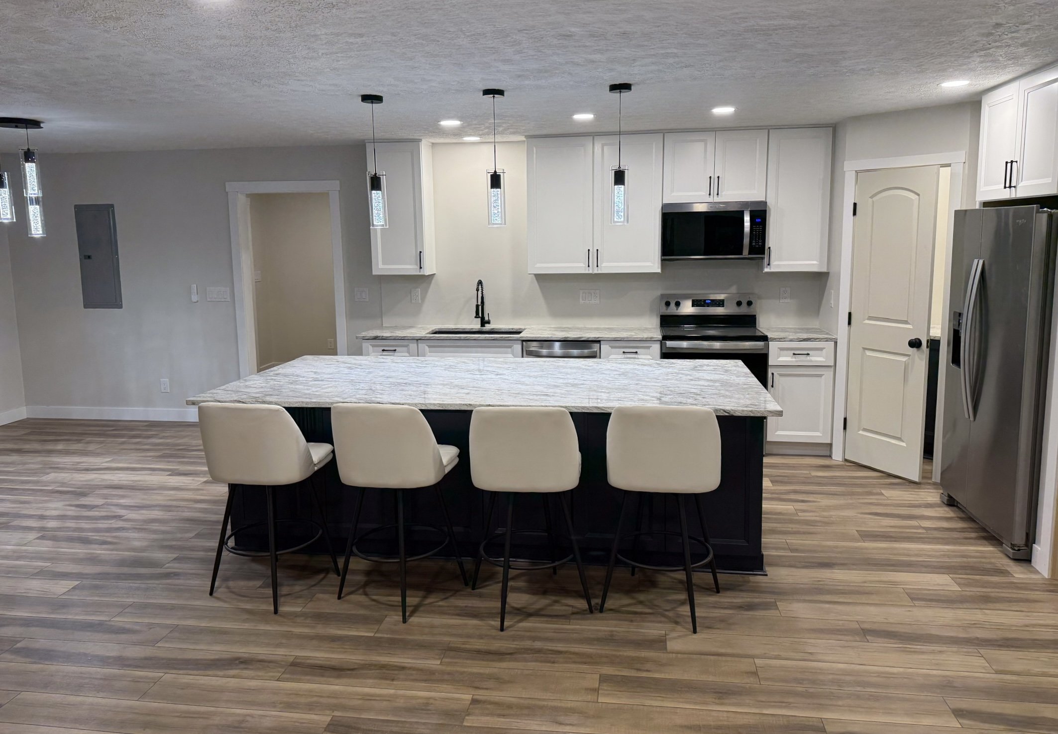 Modern, spacious kitchen and dining area with white cabinets, stainless steel appliances, a marble kitchen island, and a set of cream dining chairs, illuminated by pendant and recessed lighting, with LVP flooring.
