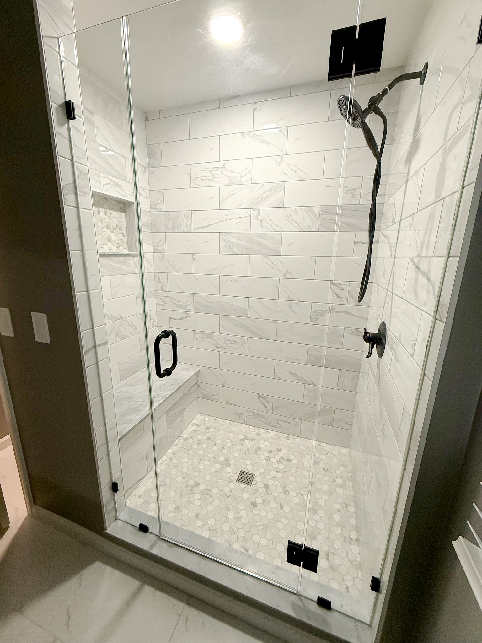 A modern walk-in shower with white subway tiles on the walls and a glass door with black hardware. There is a built-in niche with a hexagon tile pattern for shower essentials and a bench. The shower has a rainfall showerhead and a black handle.