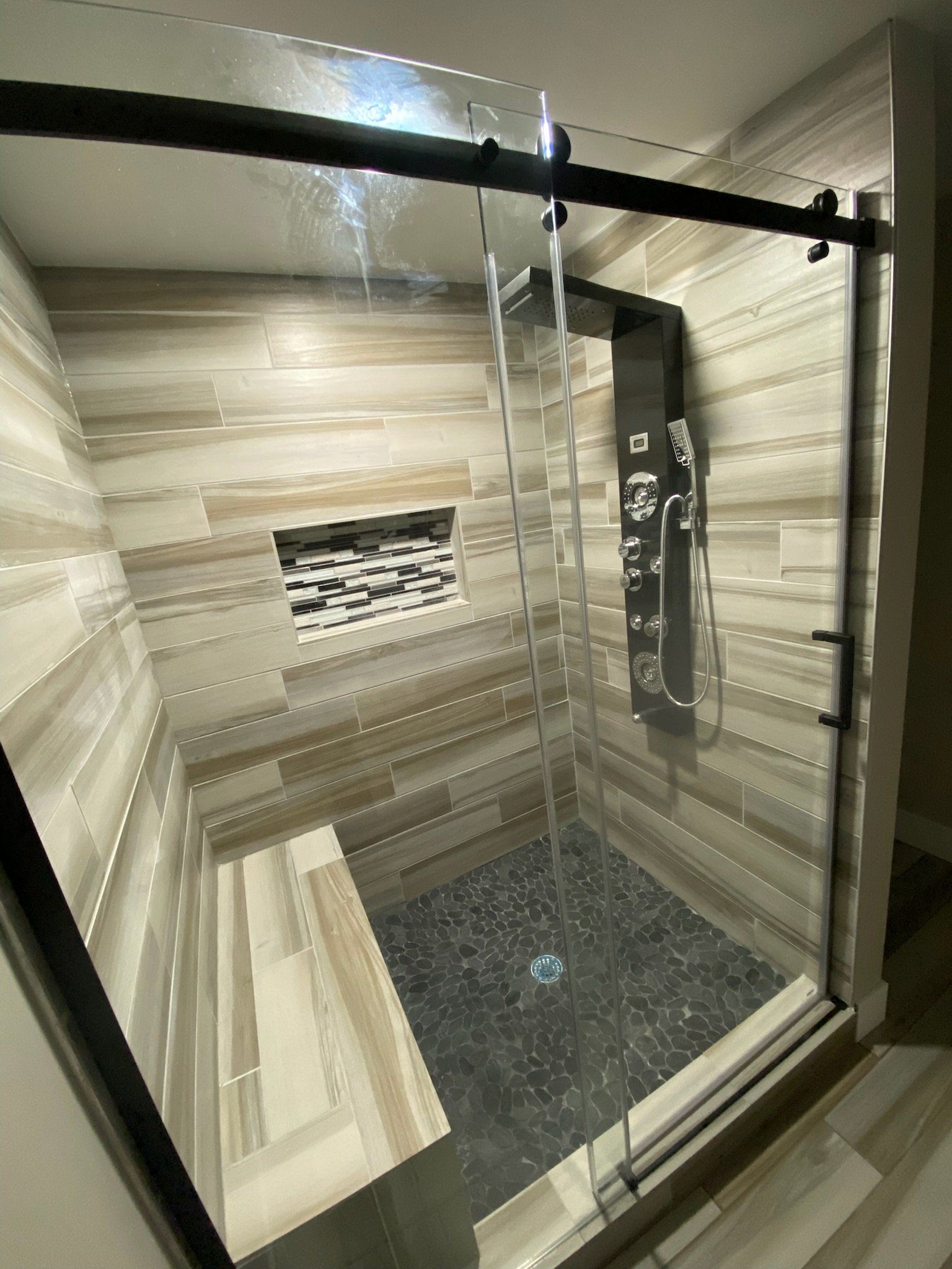 Modern shower with glass enclosure, beige and brown tiled walls, pebble floor, and built-in niche with mosaic tiles.