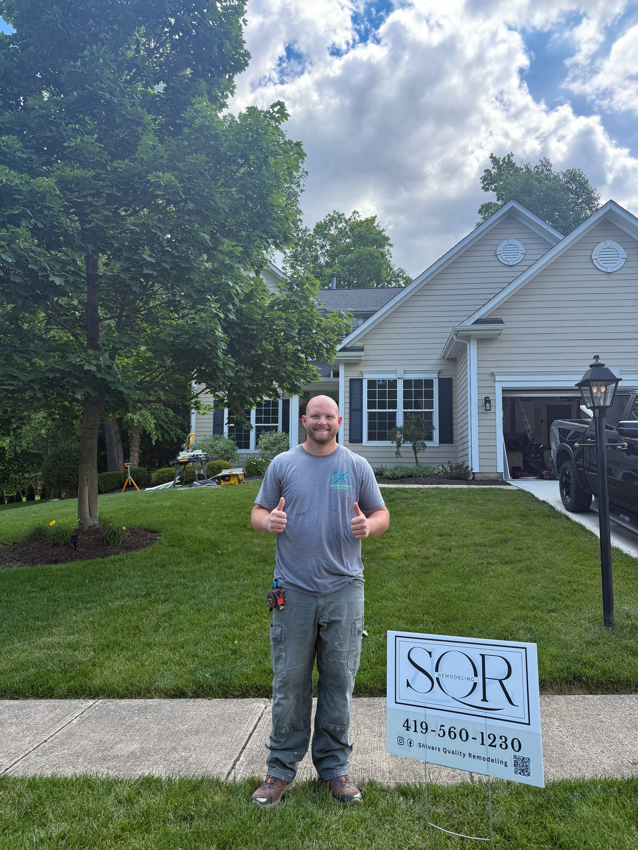 A man smiling and giving a thumbs-up in front of a house with a lawn and trees. There is a sign for a remodeling service with contact information in the grass.