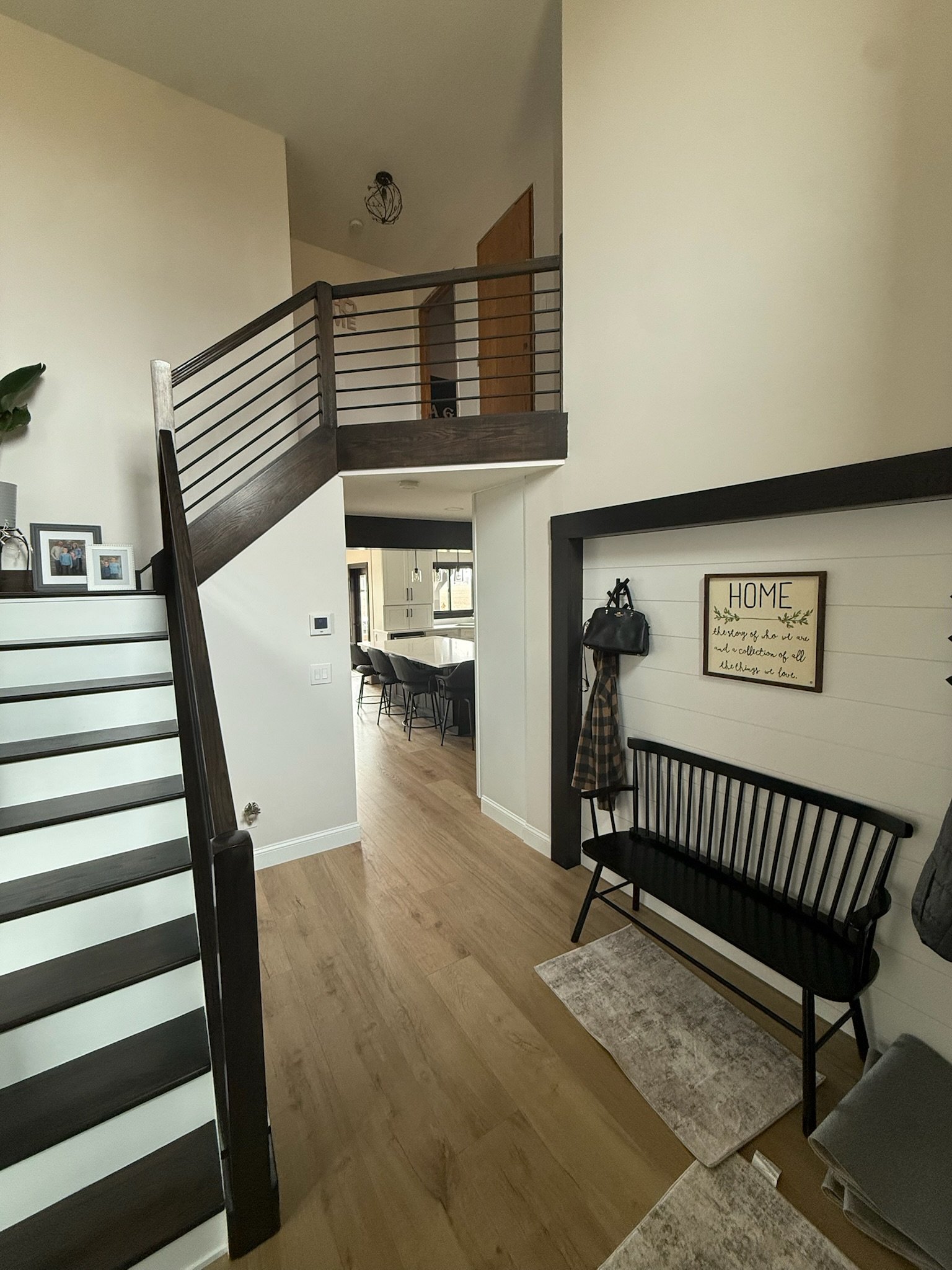 Interior of a home with stairs, stained stair railing, a dining table with chairs, a black bench, framed pictures, a wall decor with a quote, and a small rug in the entryway.