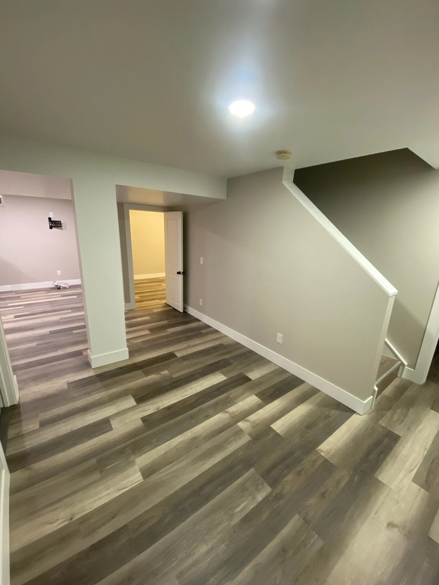 Interior view of a modern basement with wood-look flooring, light mint green walls, stairs leading upstairs, and an open doorway.