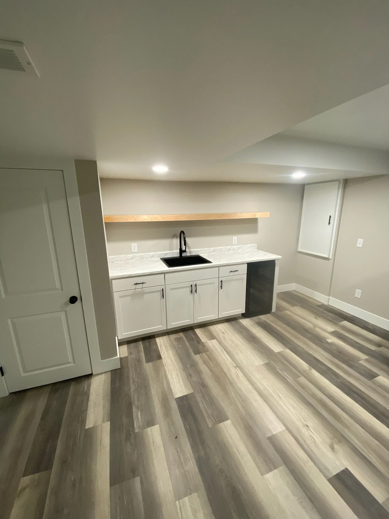 Finished basement with white cabinetry, marble countertop, black sink, wooden shelf on beige wall, and wood-look flooring.