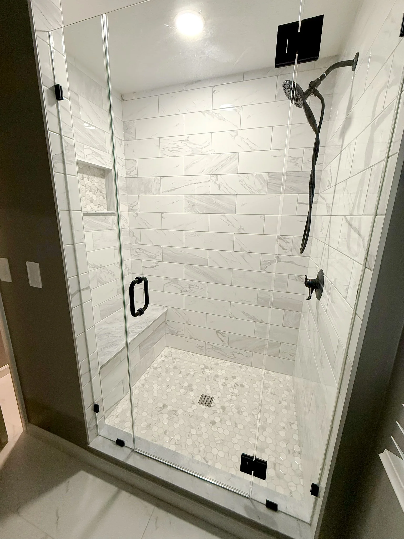 Modern glass-enclosed shower with white marble tiles, built-in shelf, and hexagonal tile floor.