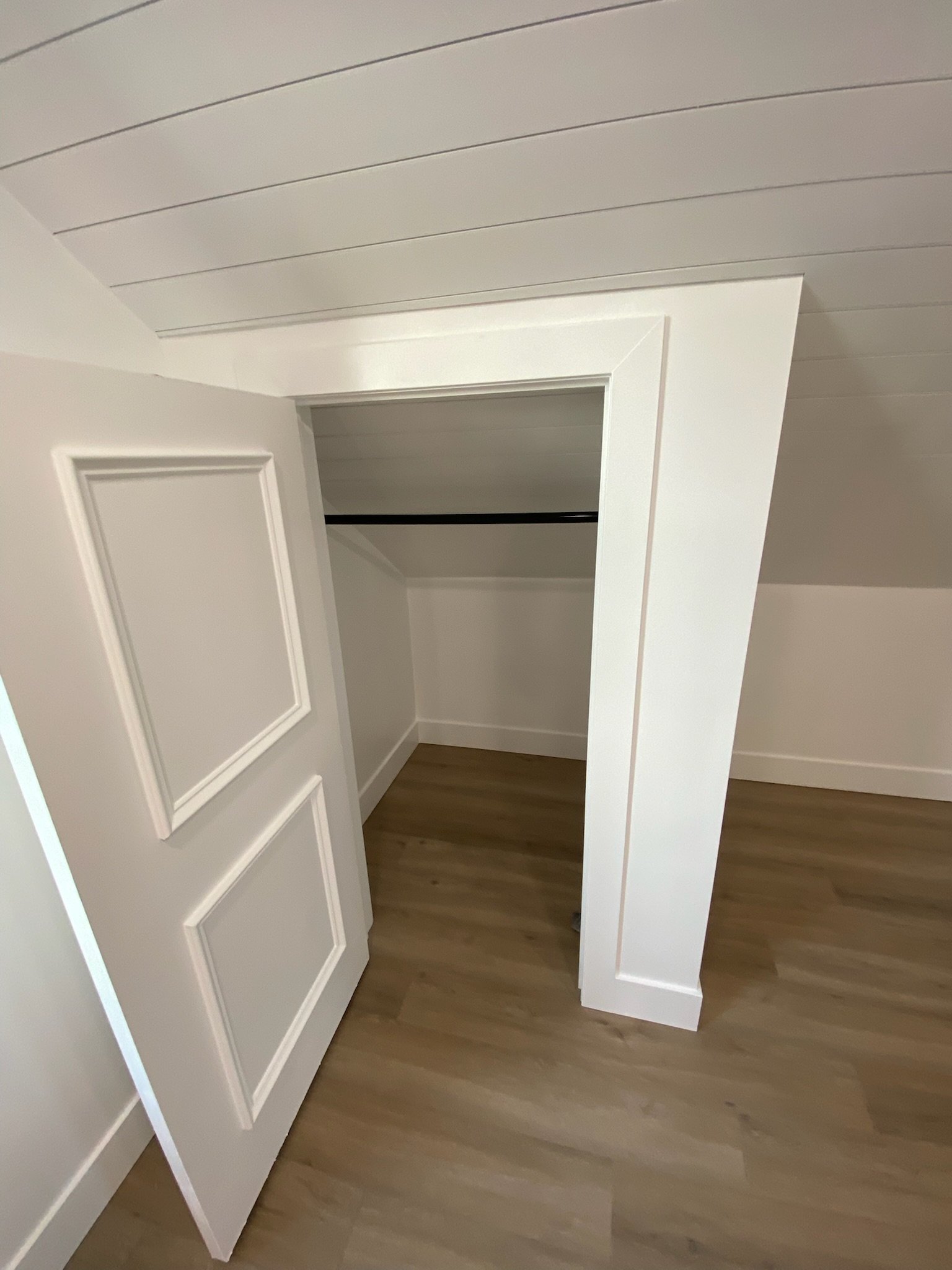 Empty closet with white walls and a white door slightly open, revealing a black hanging rod inside, and wooden flooring.