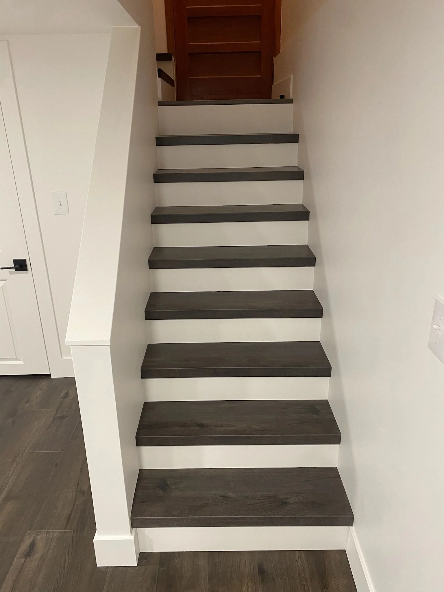 Interior staircase with dark gray steps and white sides, positioned against a white wall with a door on the left side and a doorway at the top of the stairs.