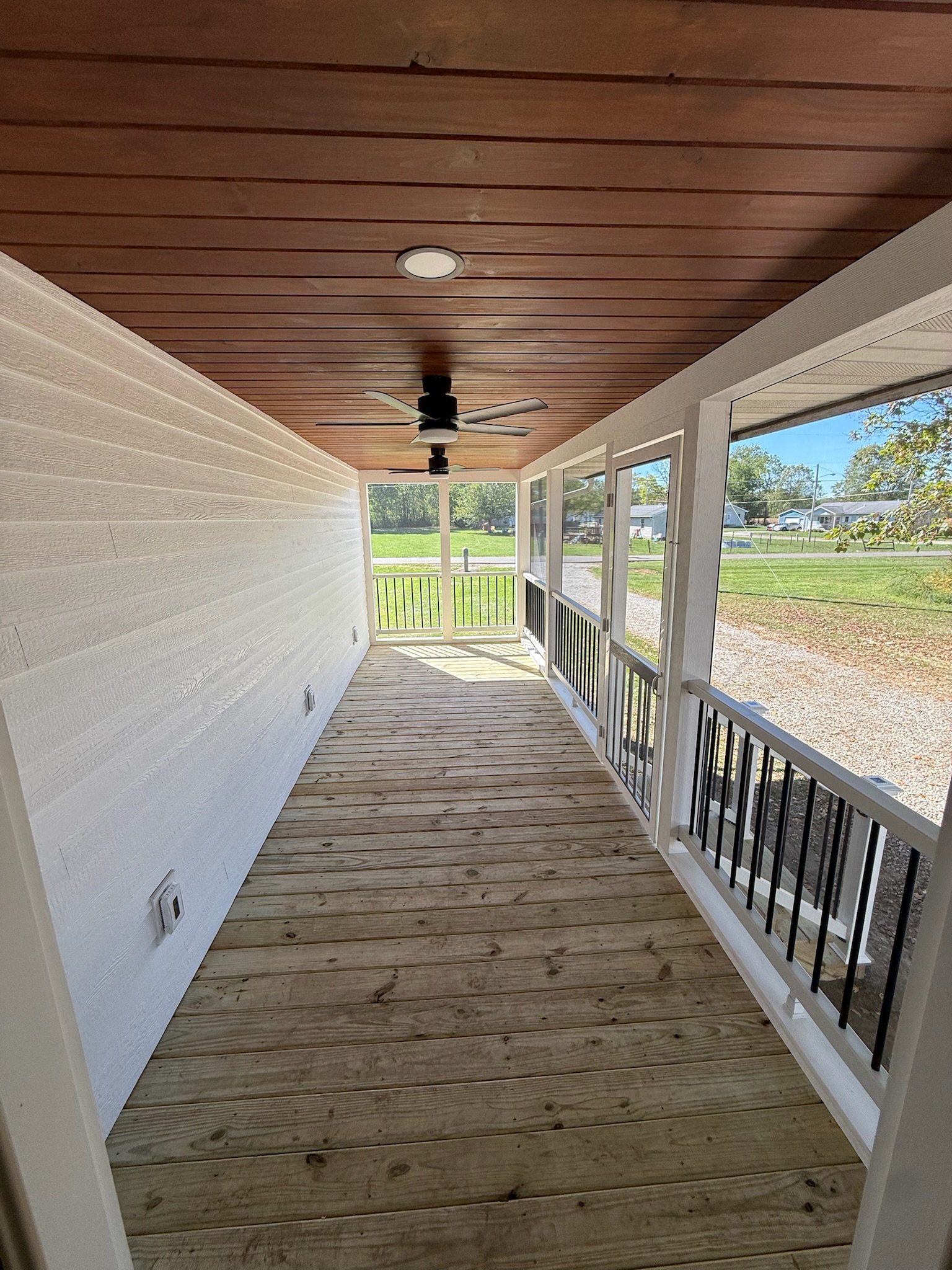 Enclosed porch with wooden ceiling, ceiling fans, white panel walls, wooden plank floor, and view of green grass and houses outside.
