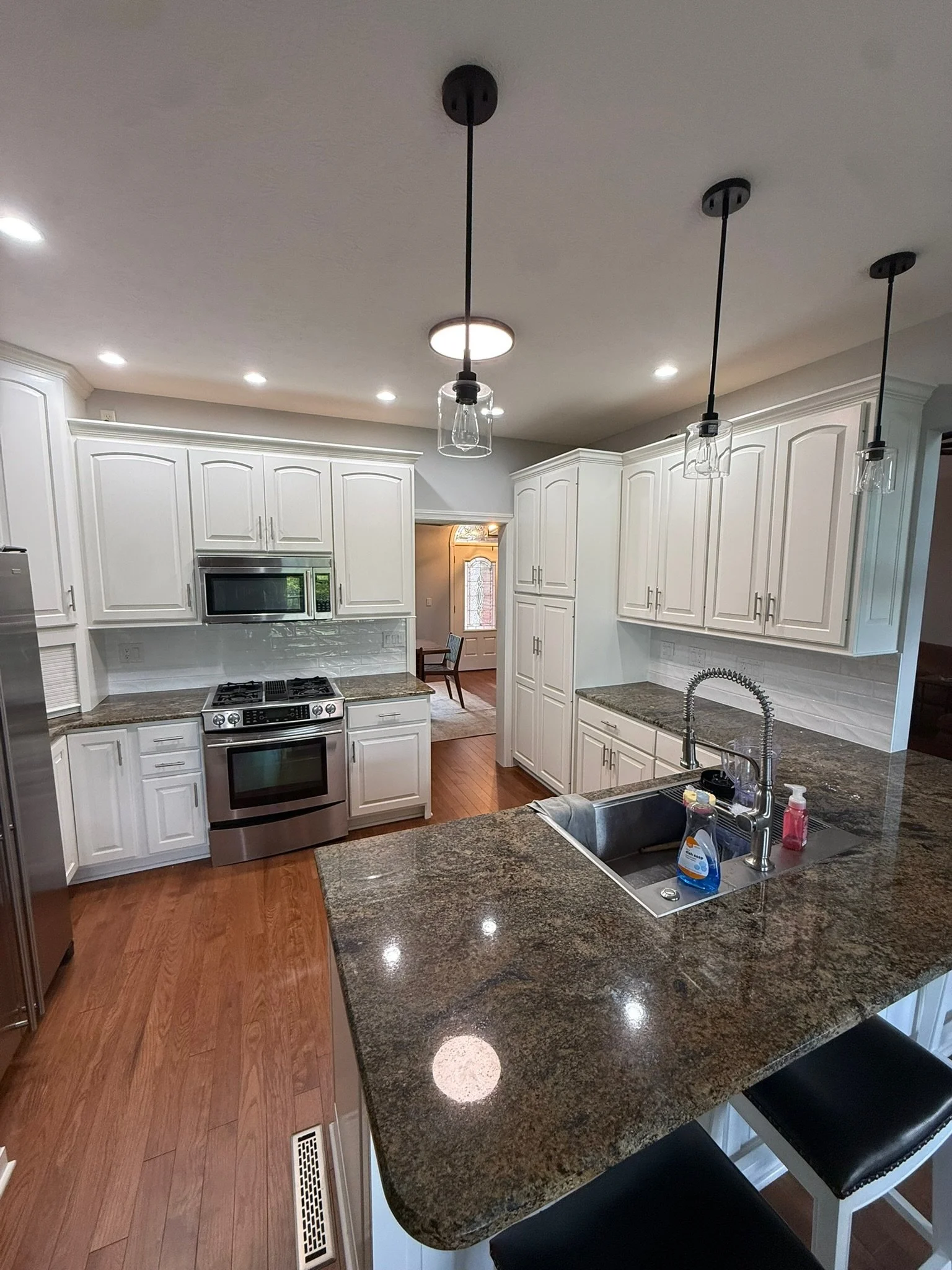 Modern kitchen with white cabinets, stainless steel appliances, granite countertops, and pendant lights. Hardwood floors, a kitchen island with bar stools, and backsplash.