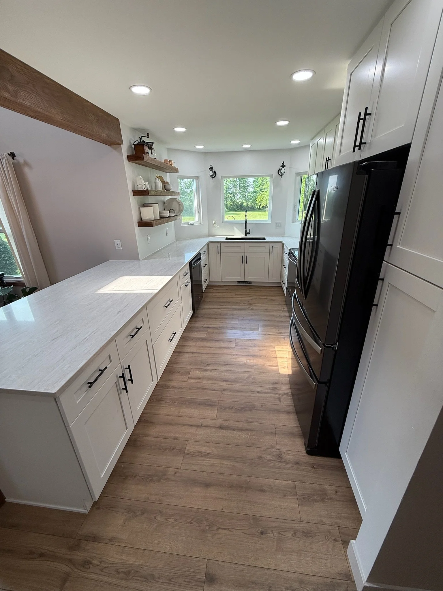Modern kitchen with white cabinets, black appliances, wooden flooring, and large windows with a green landscape outside.