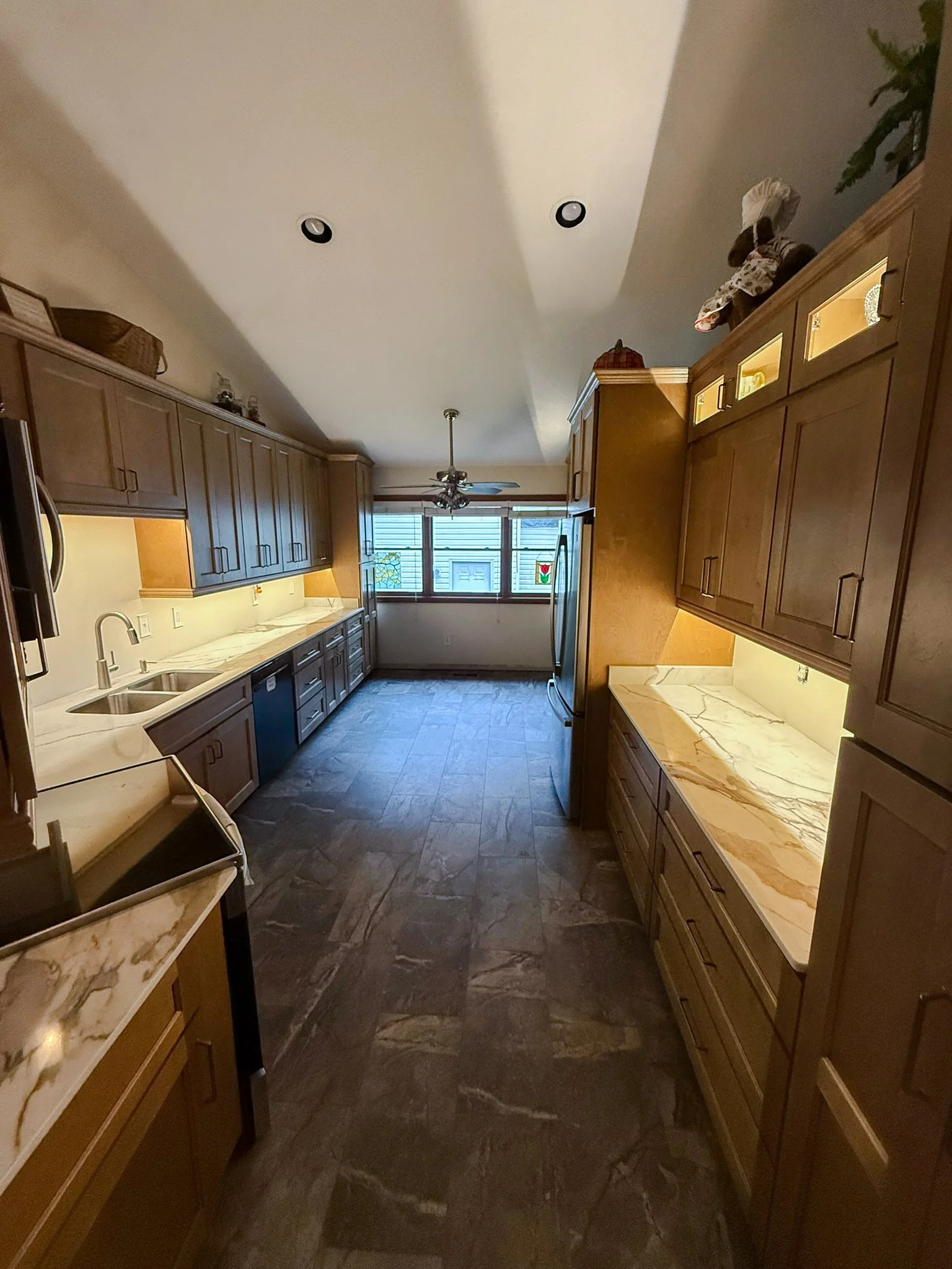 Interior view of a kitchen with brown cabinets, marble countertops, and a window, featuring a ceiling fan and recessed lighting.