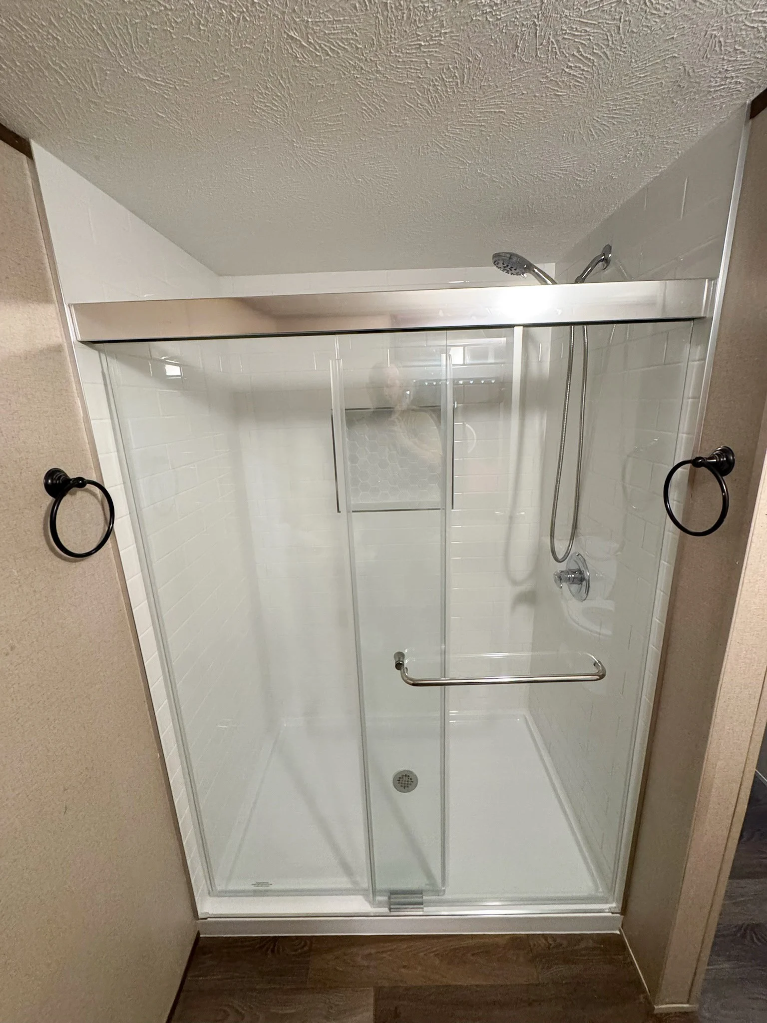 A shower with sliding glass doors, white tile walls, a showerhead, a handrail, a soap dish, and a drain on the floor, in a bathroom with beige walls and wood flooring.