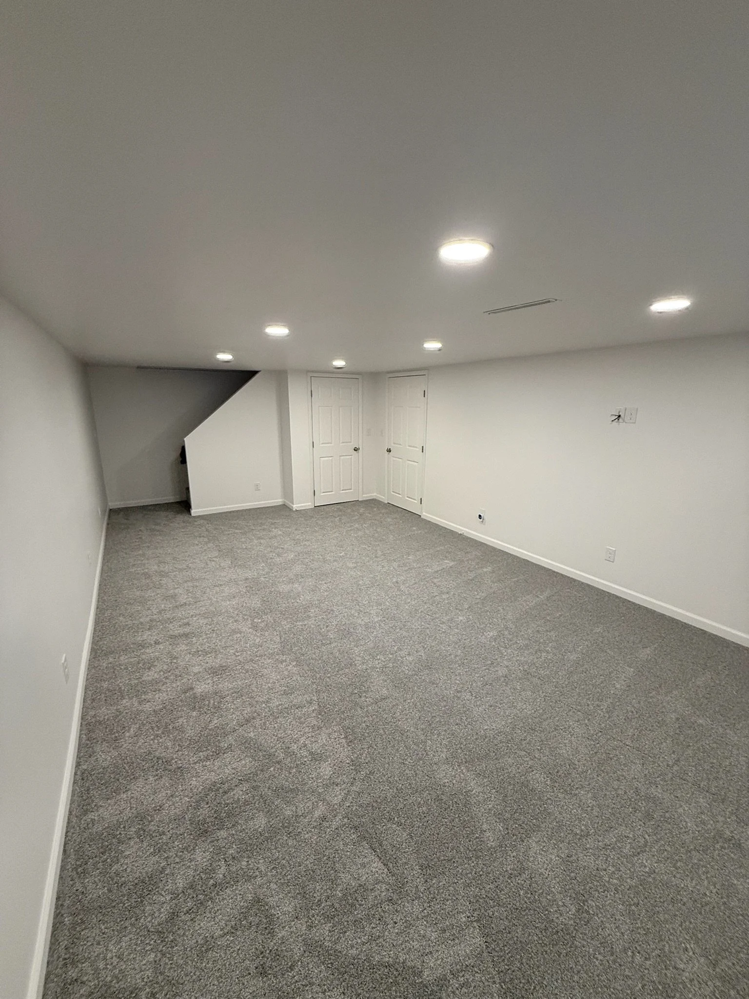 Finished basement with white walls, gray carpet, and ceiling lights, with two closed white doors and a small staircase in the corner.