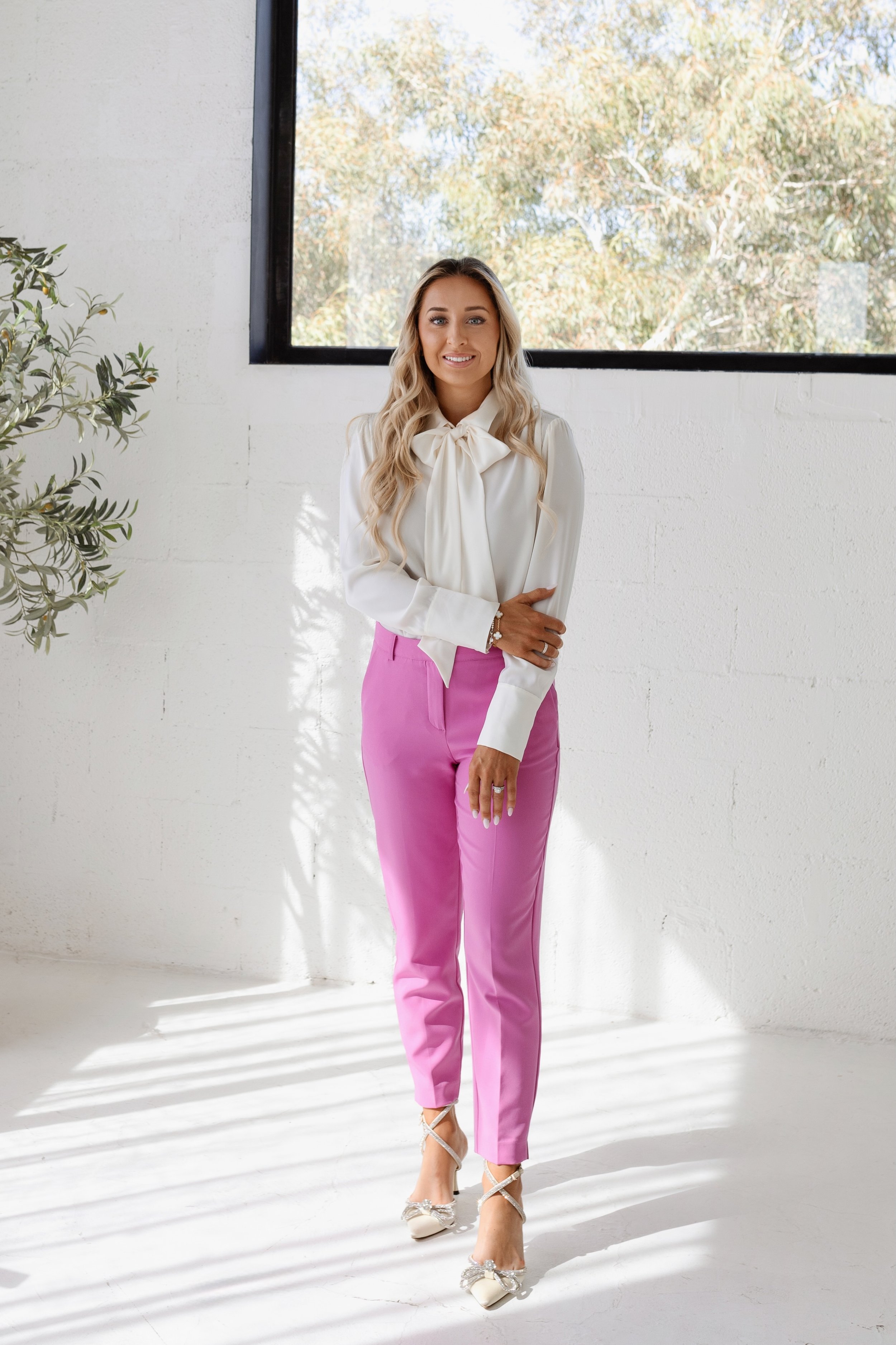 A woman wearing a white blouse with a bow tie at the neck and pink pants standing inside near a window with trees outside.