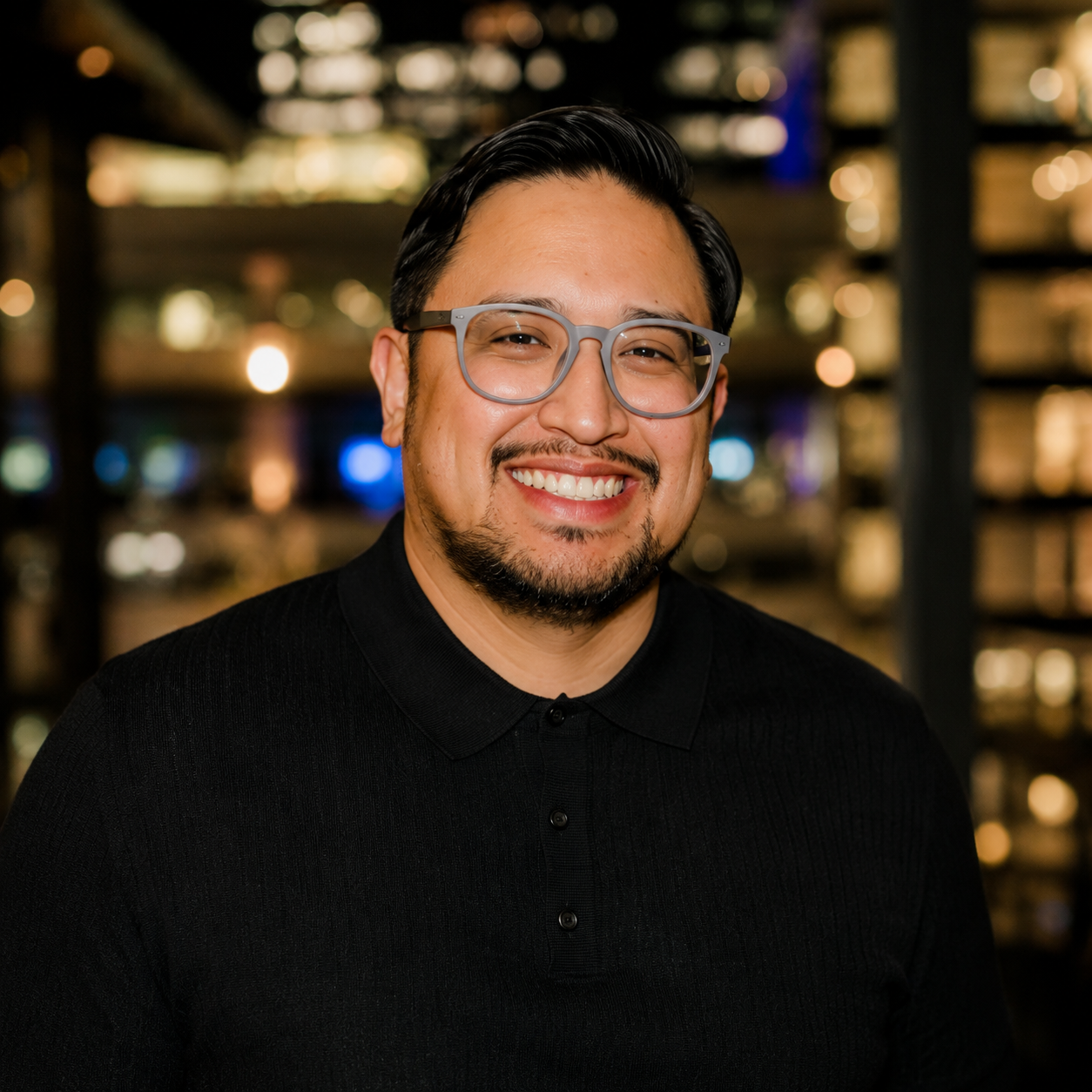 A smiling man with glasses and a goatee wearing a black shirt, standing in front of a blurred background of city lights at night.