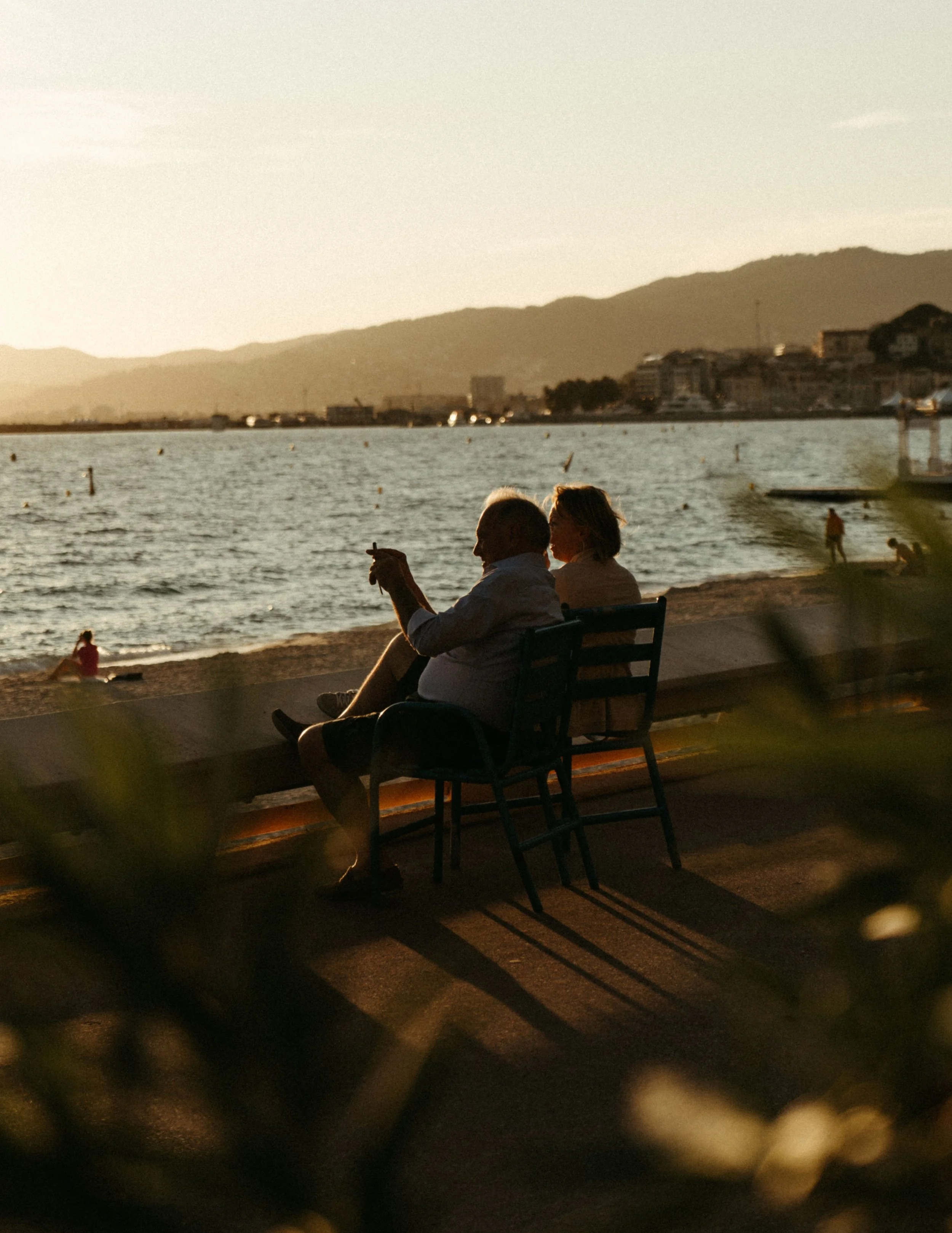 People sitting on a bench by the water during sunset, with mountains and buildings in the background.