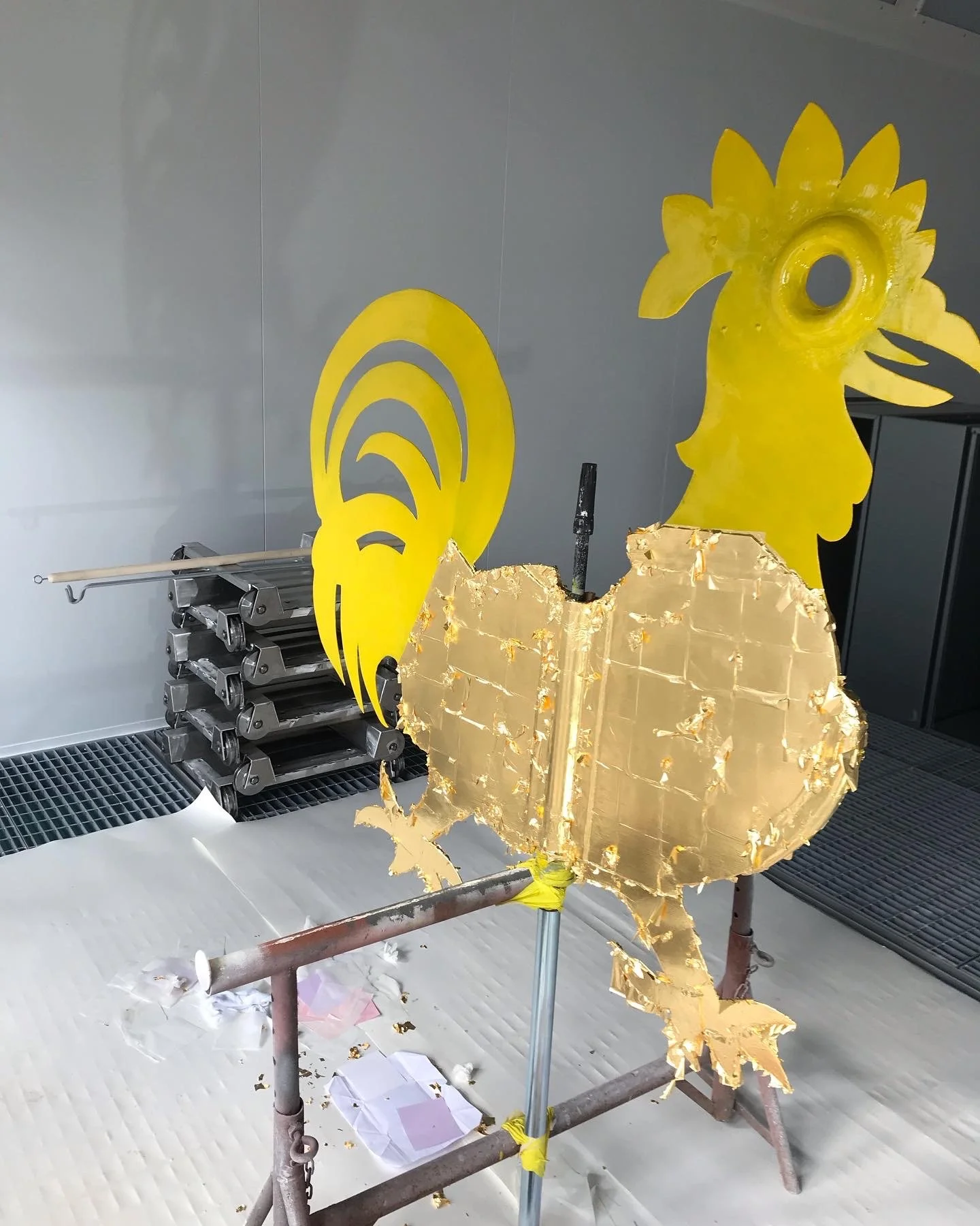 Entstehende goldene Skulptur in Form eines Hahns mit einem roten Standfuß, im Hintergrund eine gelbe Skulptur mit Bogenmustern, in einem Werkraum.