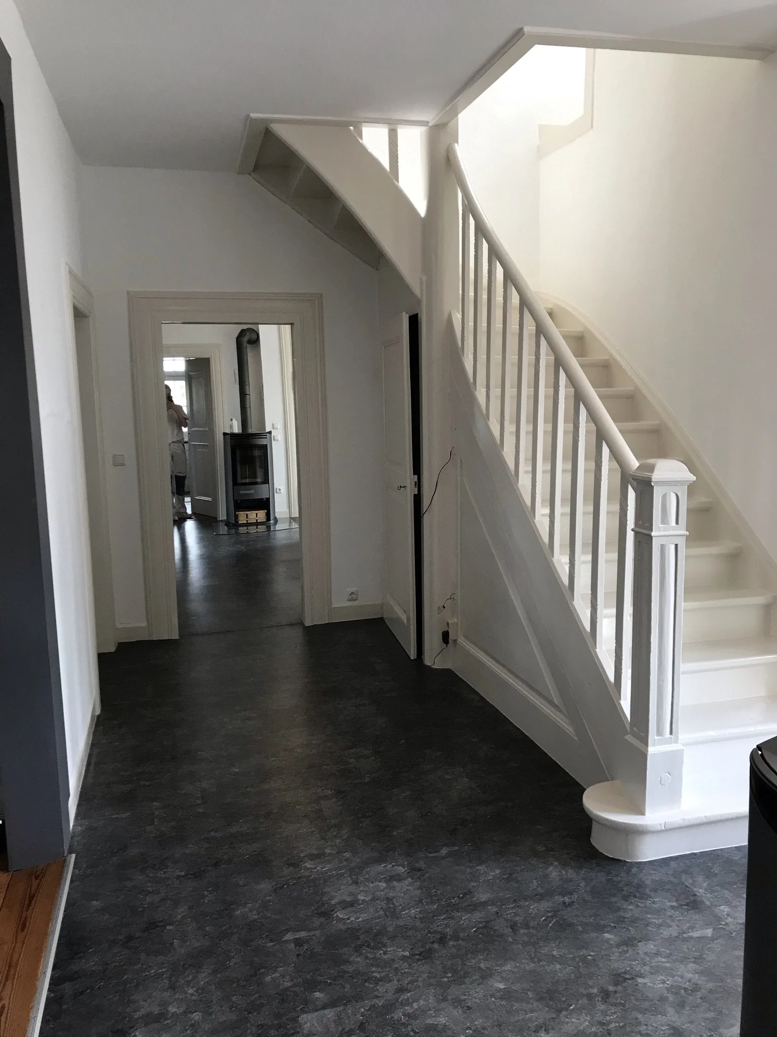 Ein Flur im Haus mit dunklem Fußboden und weißem Treppenhaus. Im Hintergrund ist eine Tür und ein Kaminofen im Raum sichtbar.