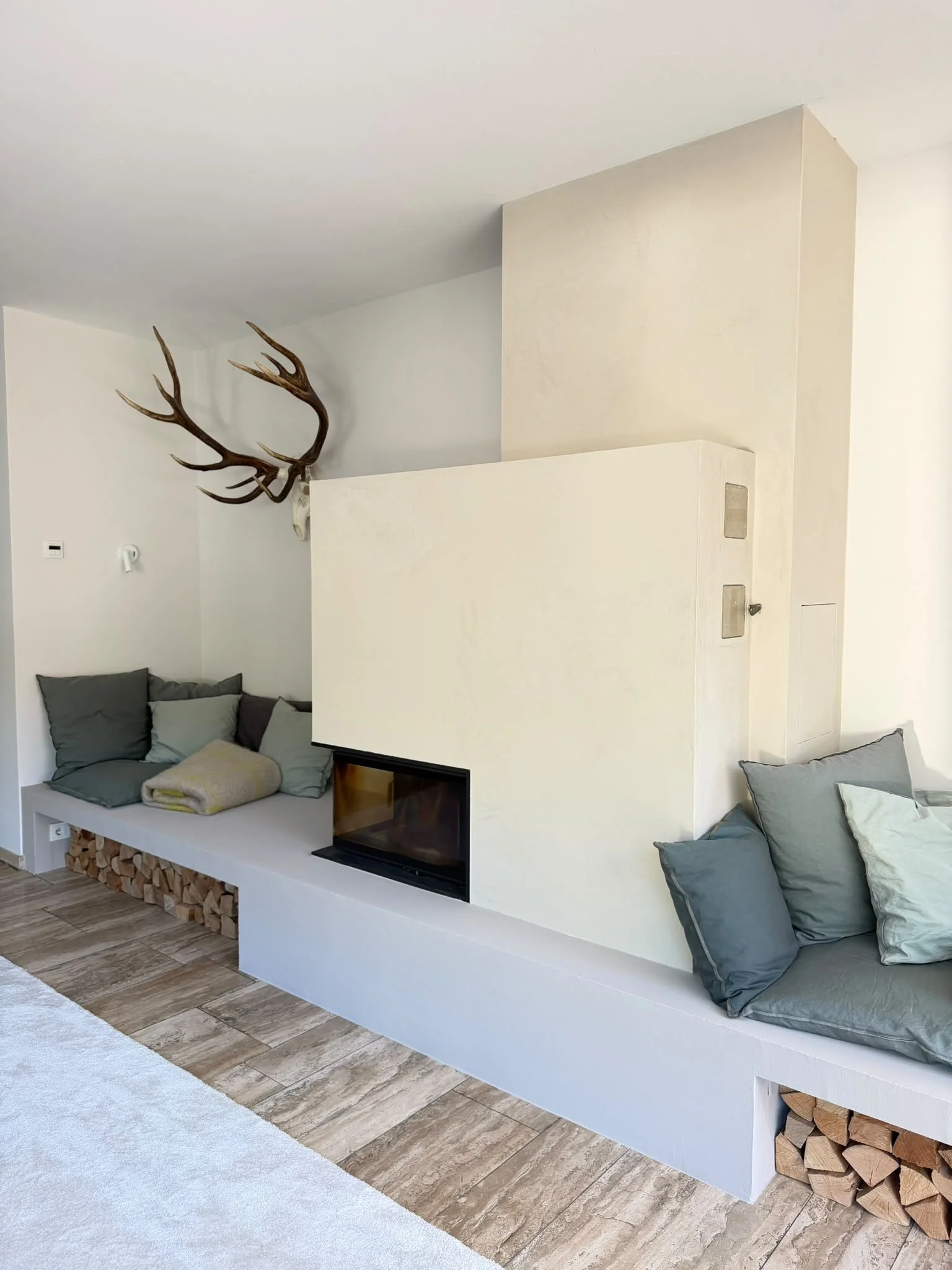 Ein Wohnzimmer mit einem weißen Sitzbereich, mehreren grauen Kissen und einem Holzfeuer im Kamin. An der Wand hängt ein Geweih. Der Boden ist mit holzähnlichen Fliesen ausgelegt.