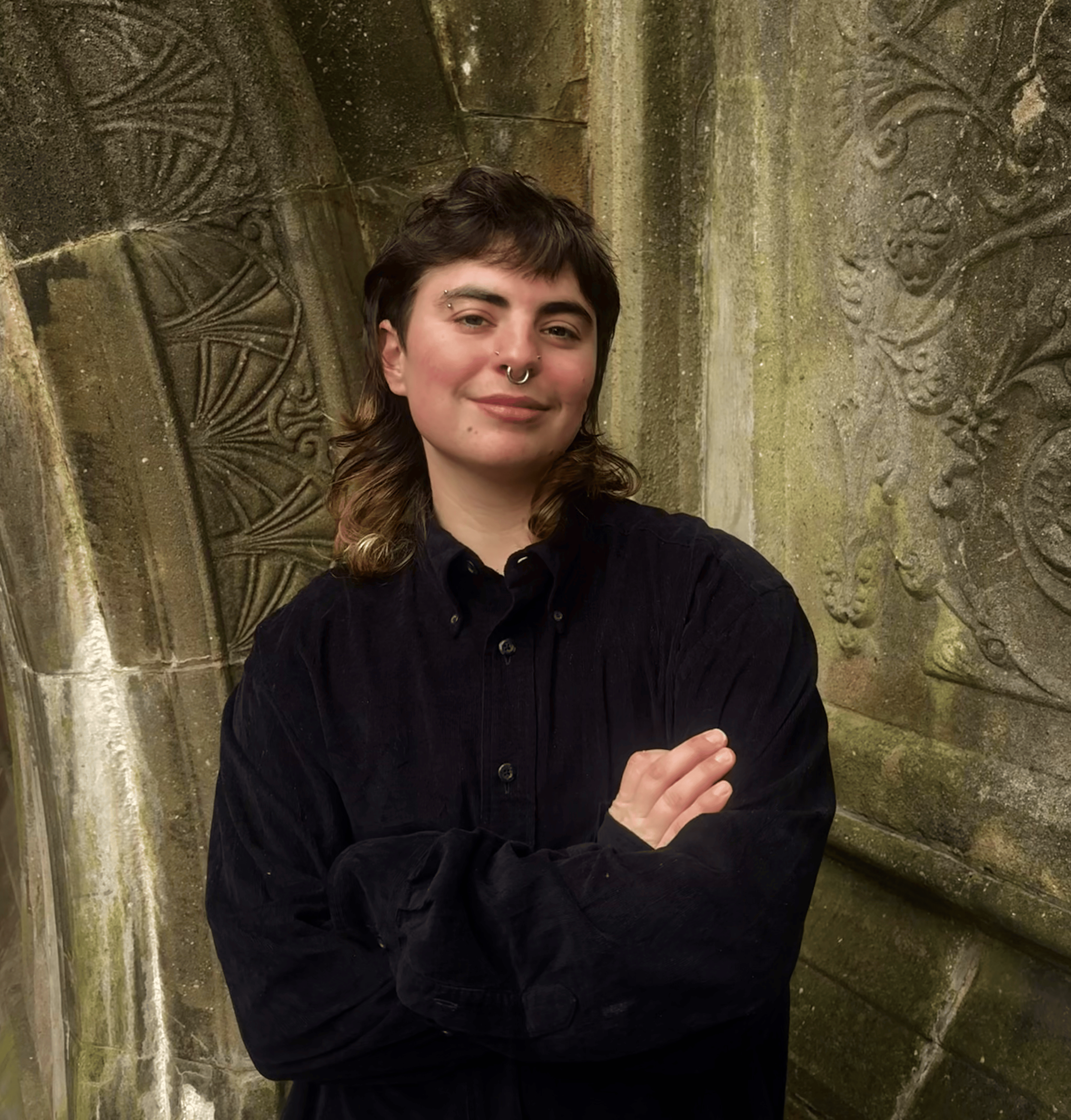 A young person with dark hair and a nose piercing, wearing a black shirt, standing against a stone wall with carved designs.