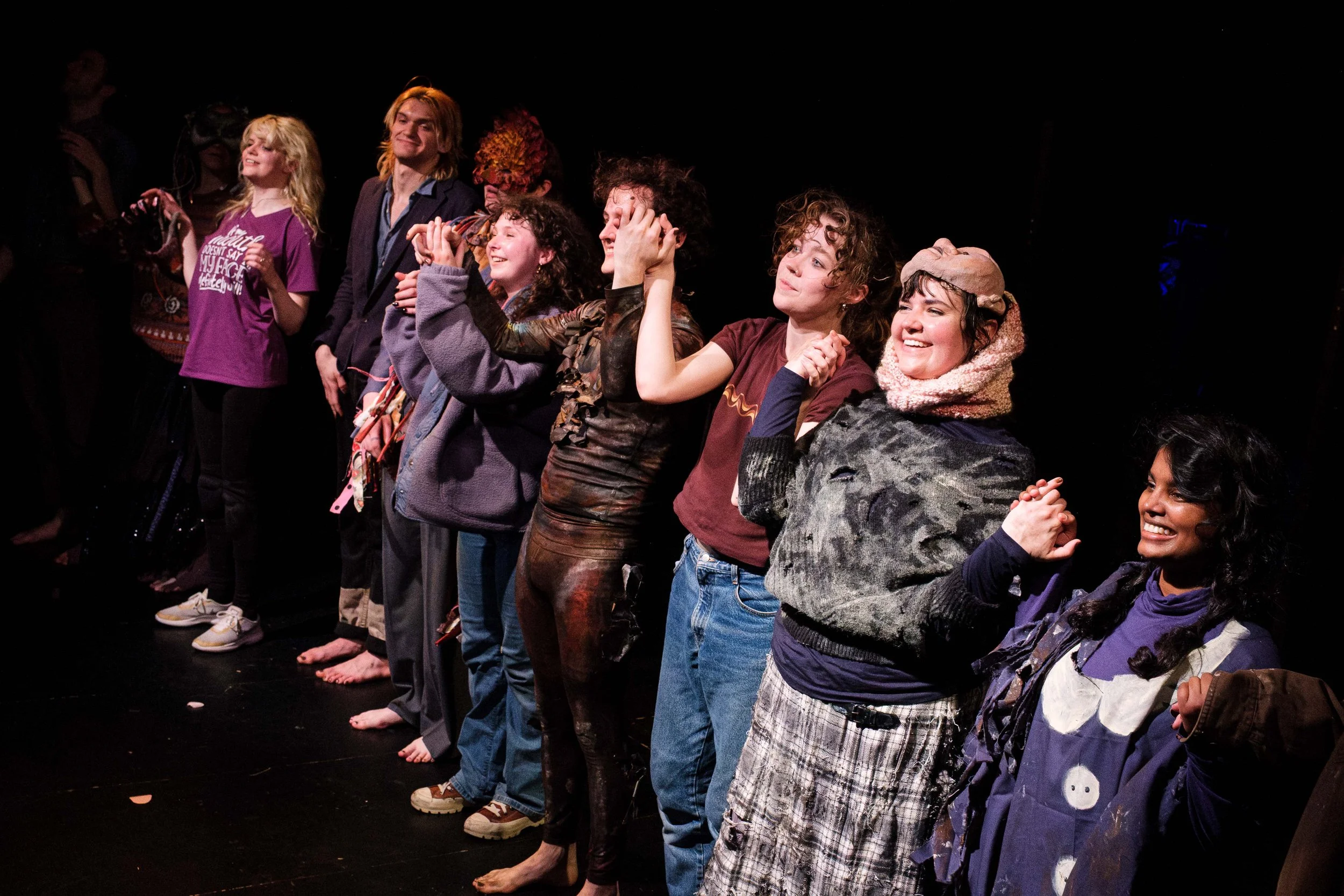 Group of people standing in a line on a stage, smiling and reacting to a performance, some with hands on their heads, dressed casually, with a dark background.