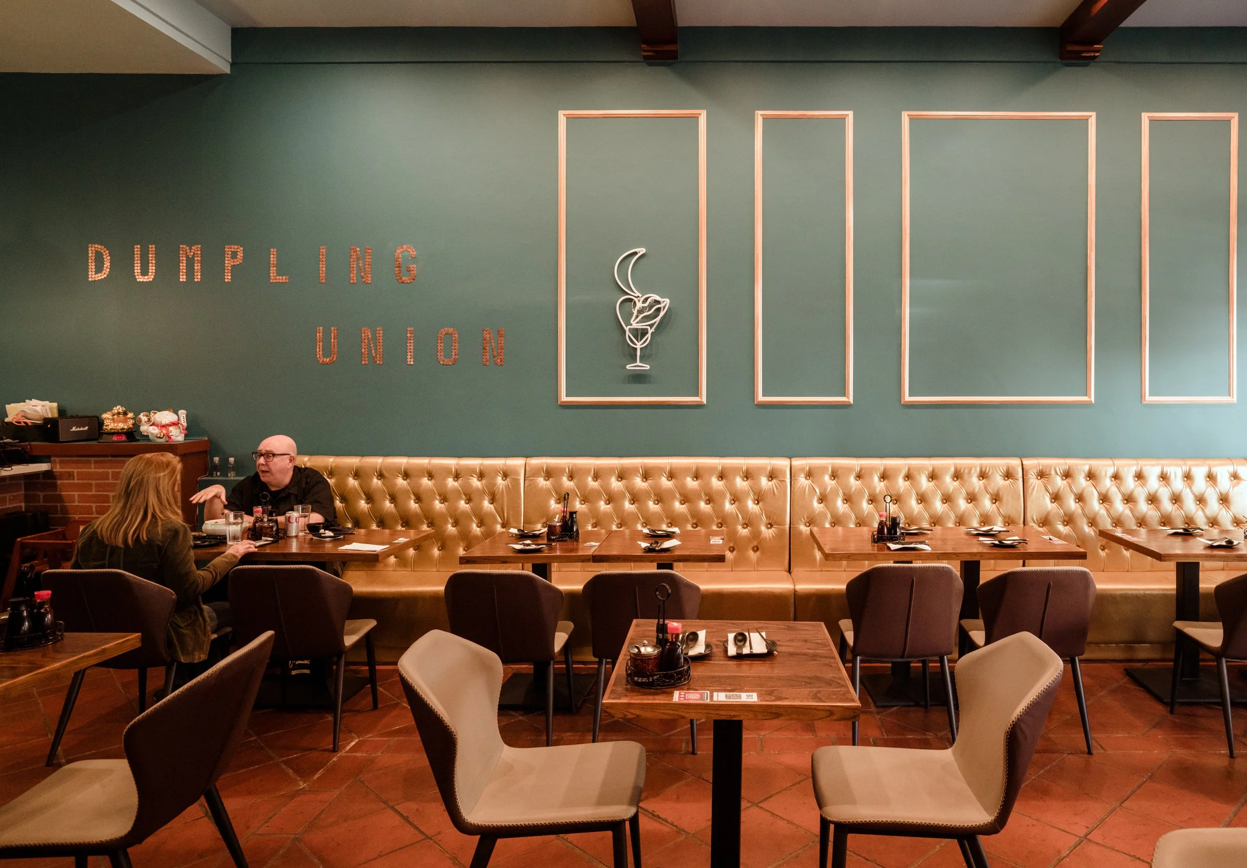 Inside a restaurant with a green wall, 'DUMPING UNION' spelled out on the wall, a neon sign of a cocktail glass with a crescent moon, and tables set for dining with two people engaged in conversation.