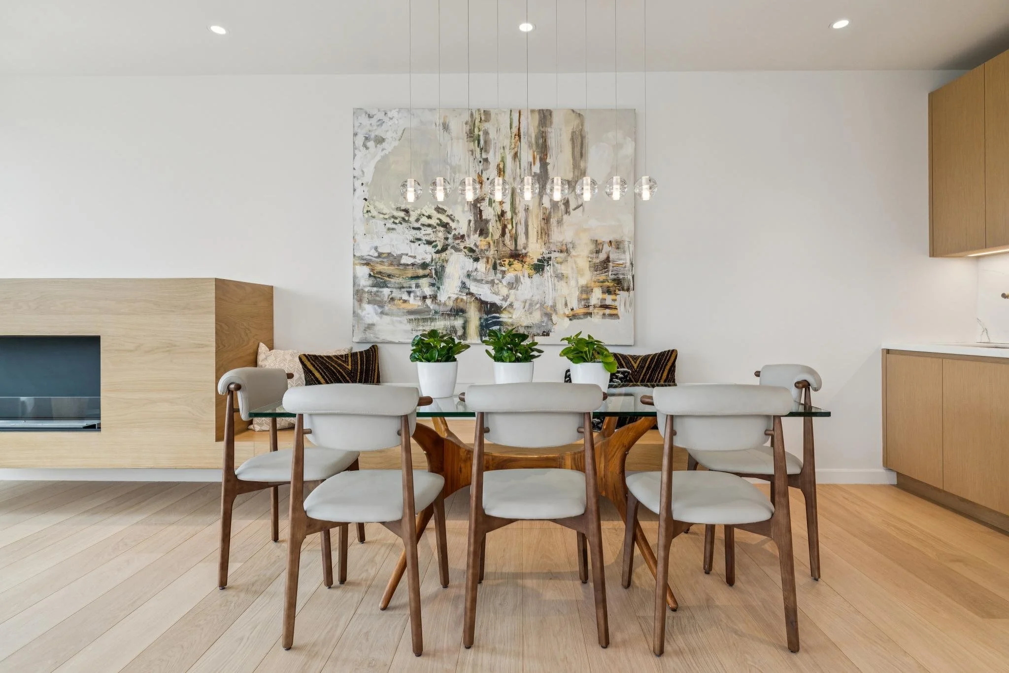 Modern dining room with a glass-top table, eight white upholstered chairs with wooden legs, a large abstract painting on the wall, and three green potted plants on the table, with a wooden cabinet in the corner.