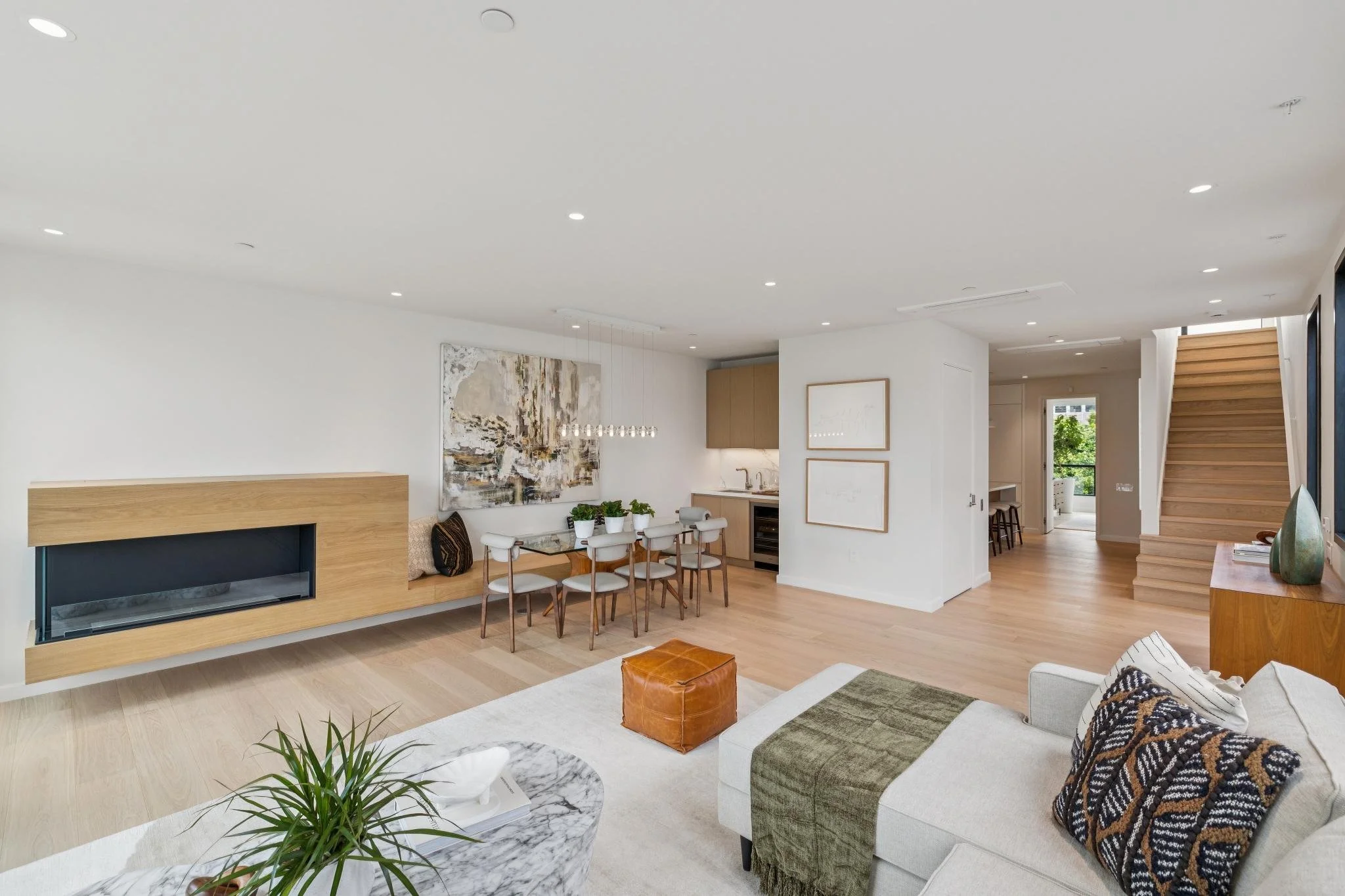 Interior of a modern, minimalist living room with white walls, hardwood floors, a built-in fireplace, a dining area with chairs, artwork, and a staircase.