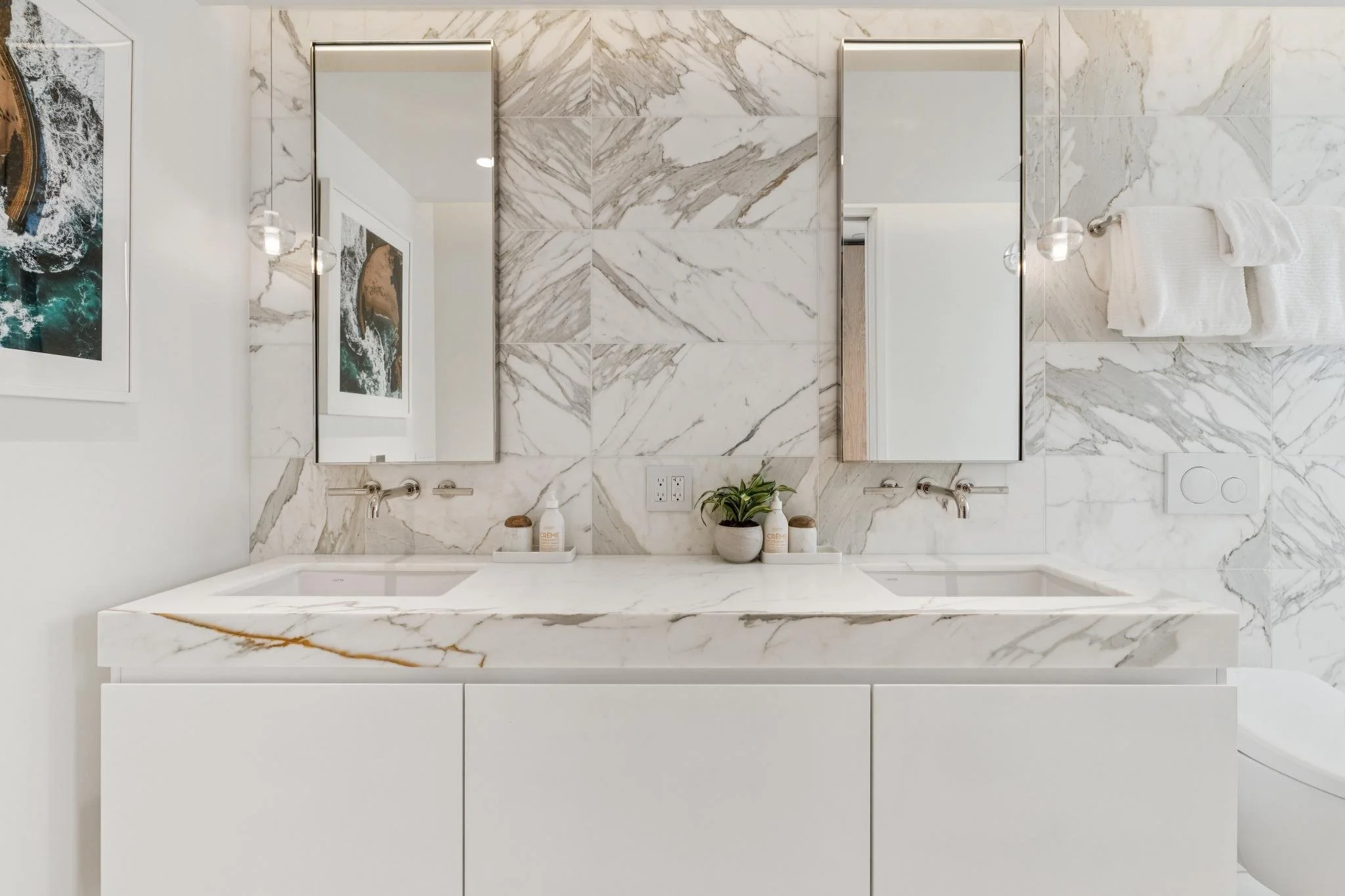 A modern bathroom with a white marble double vanity, two large mirrors, and minimalist decor including small plants and toiletries.