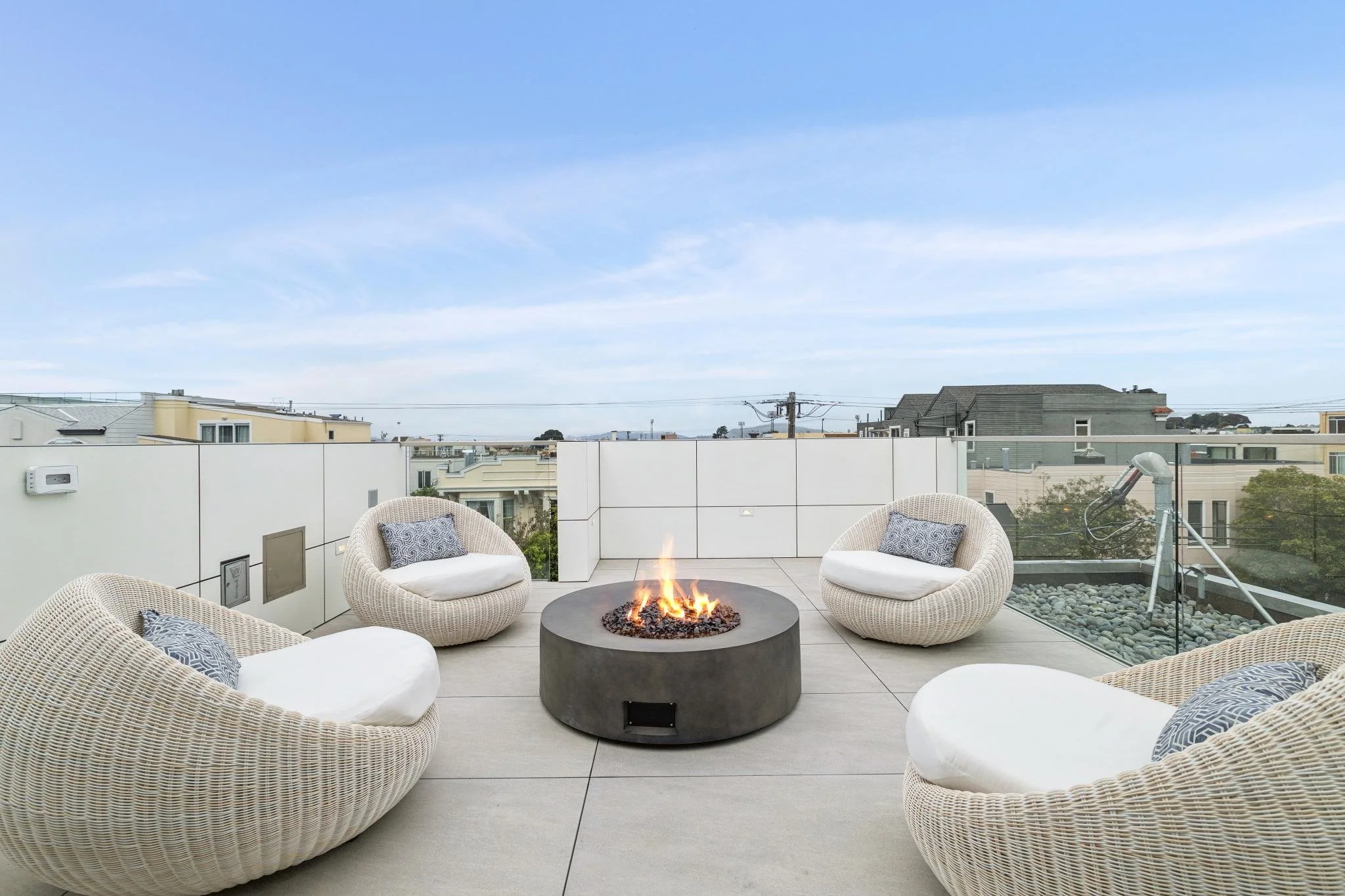 Outdoor rooftop patio with wicker chairs, pillows, and a fire pit, overlooking neighboring houses under a clear blue sky.