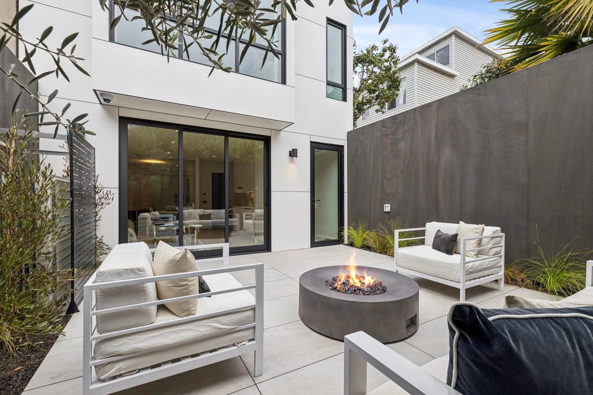 Modern backyard patio with white outdoor sofas, a circular fire pit in the center, and a dark gray privacy wall. Glass sliding doors lead into the house, which has a white exterior with black window frames. There are trees and plants around the patio