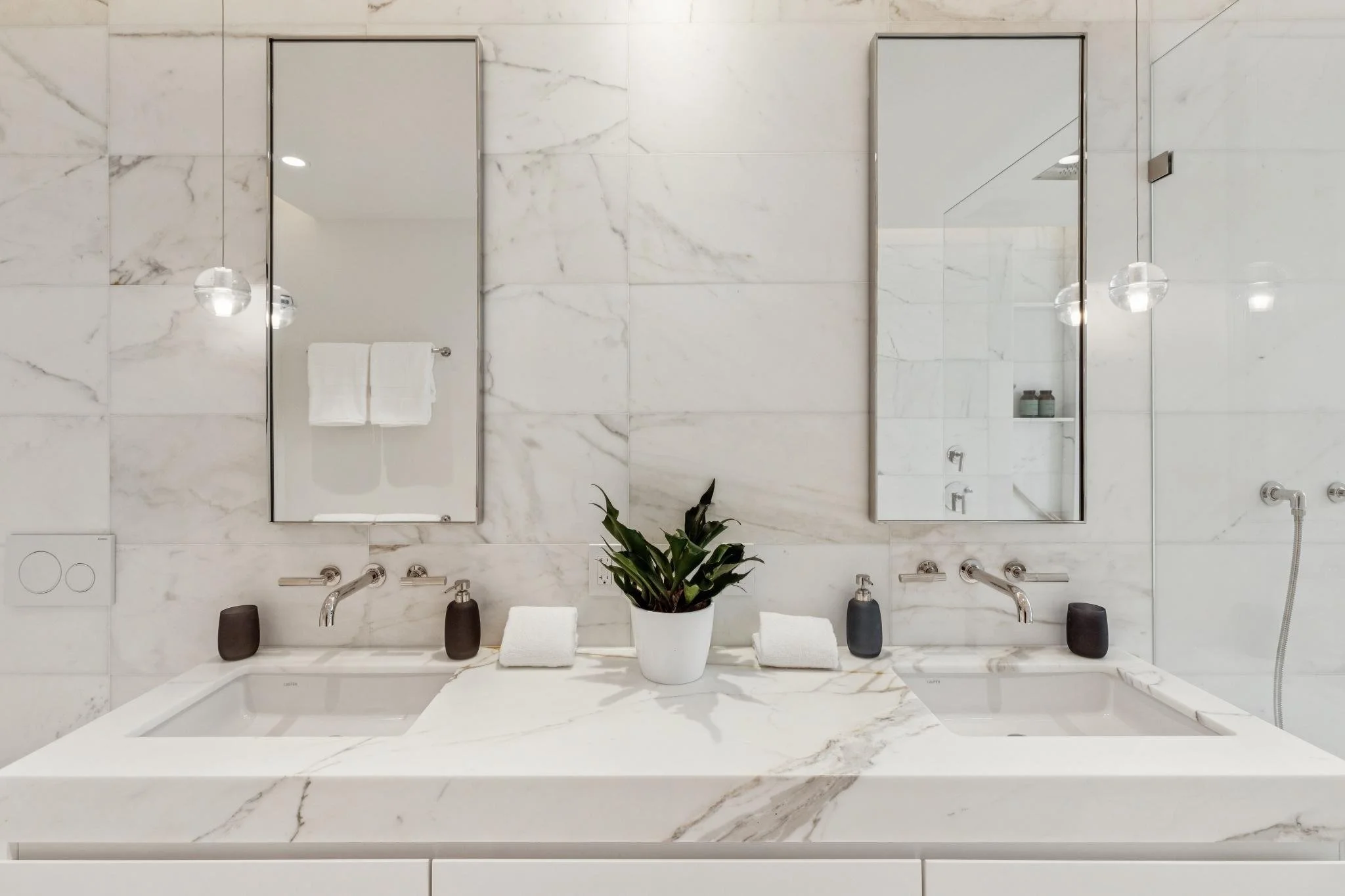 A modern bathroom with white marble walls, two large rectangular mirrors, a white sink, and a potted plant in the center.