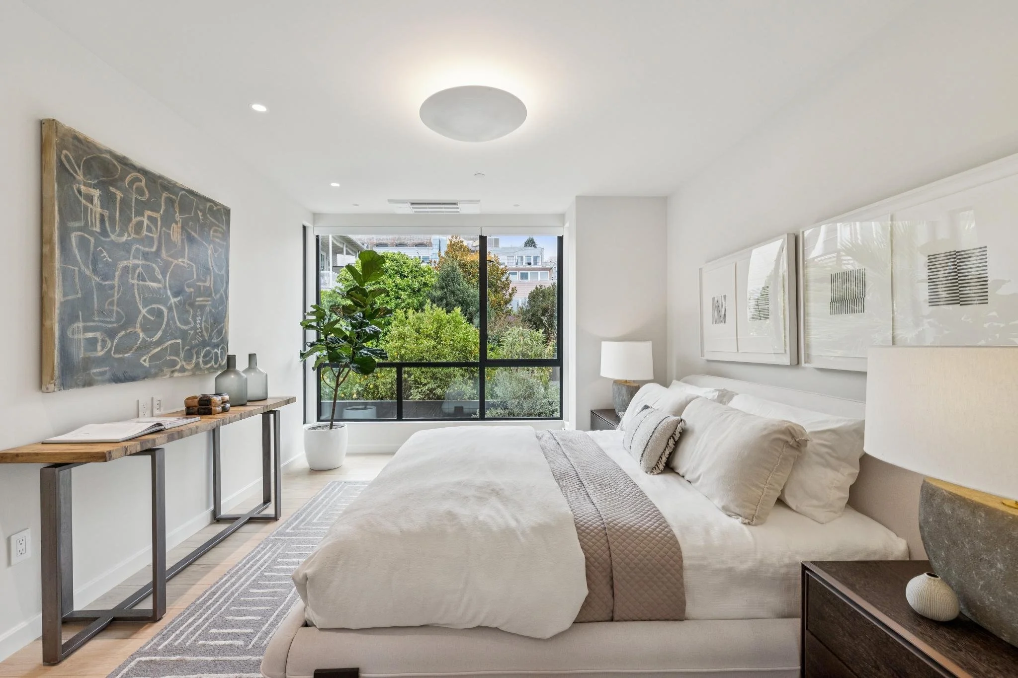 Modern bedroom with a large bed, artwork on the wall, a window overlooking greenery, a tall indoor plant, nightstand, table lamp, and decorative items.