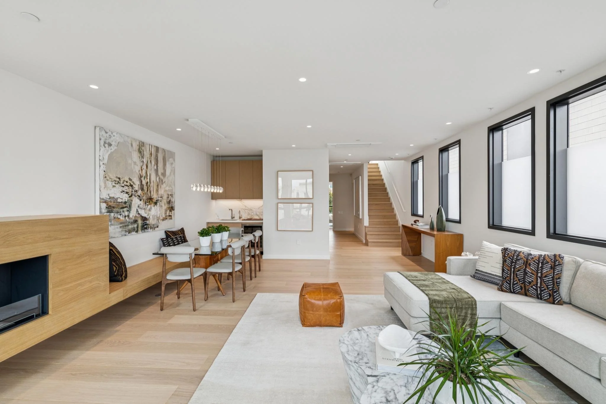 Contemporary living room with white walls, large windows, wooden flooring, a white sofa with patterned pillows, a marble coffee table with a green plant, and a wooden accent table near the staircase.