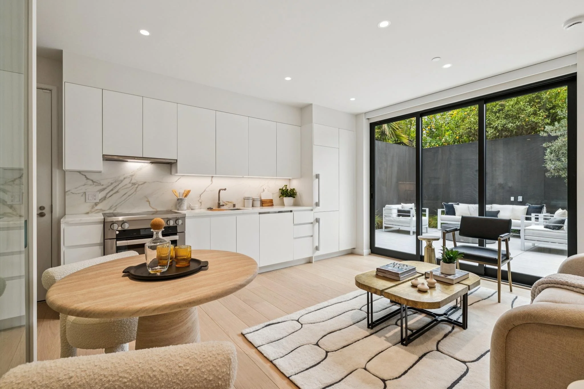 Modern kitchen and living room area with large sliding glass doors opening to outdoor patio with white seating and greenery.